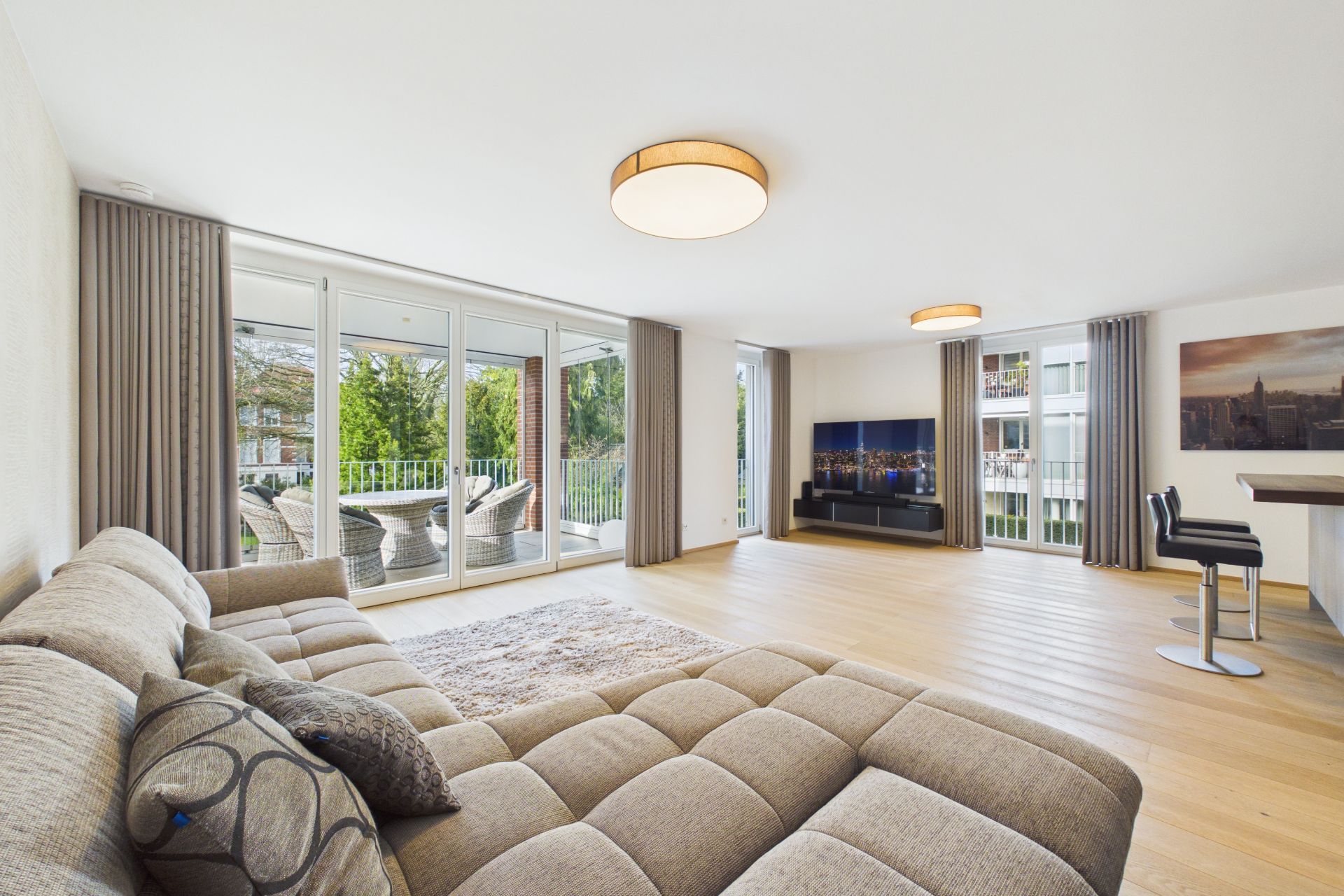 Helles Wohnzimmer mit großer Fensterfront, Ecksofa, Teppich, TV und Essbereich mit zwei Stühlen.