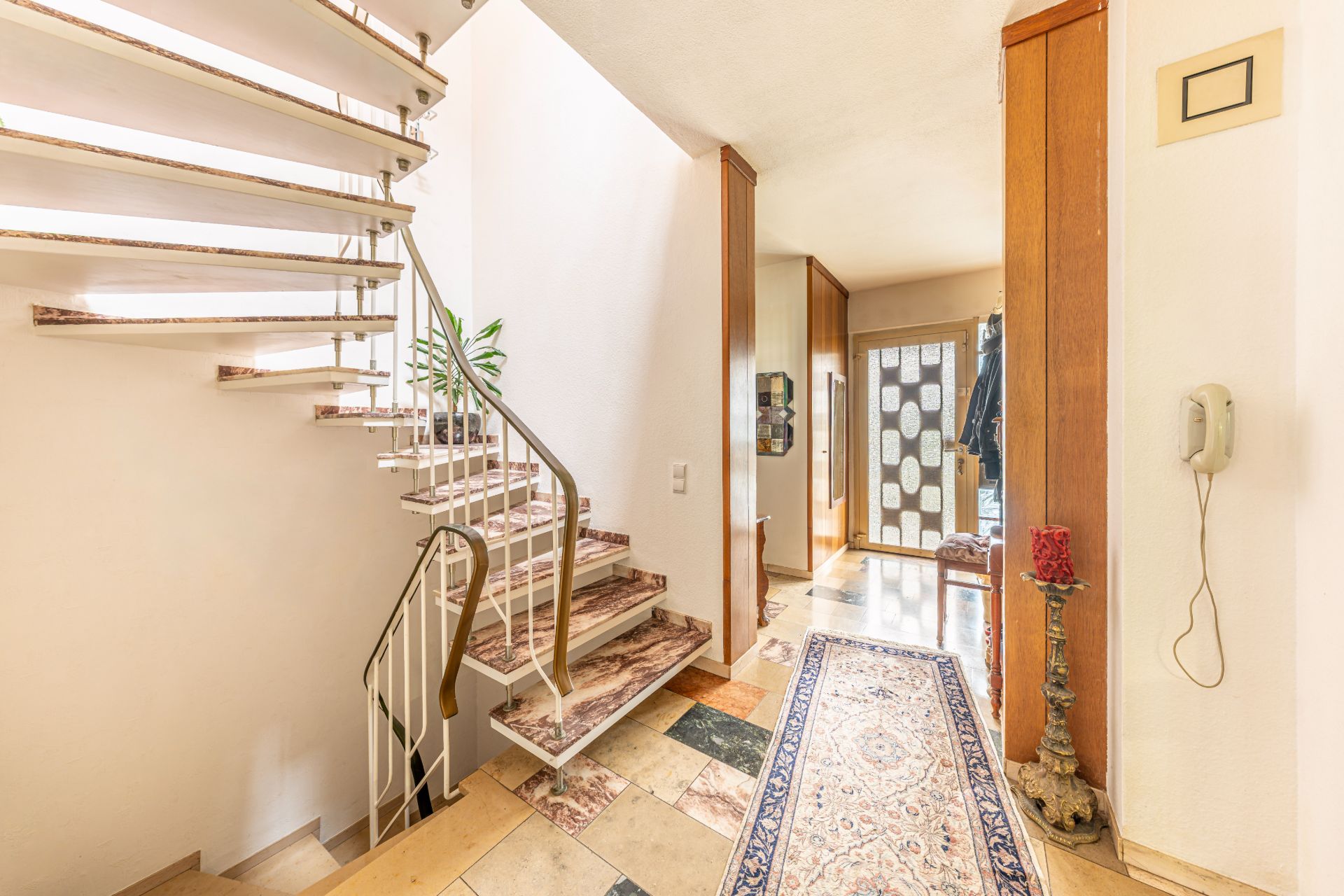 Heller Flur mit Marmortreppe, Teppichläufer und Wandtelefon in einem Wohnhaus.