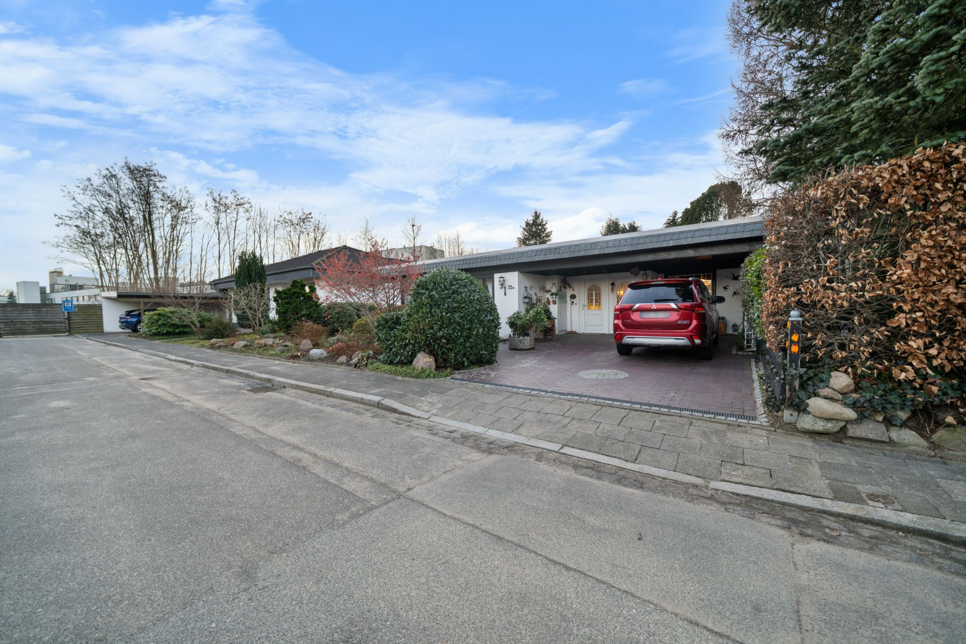 Einfamilienhaus mit Carport und rotem Auto, umgeben von Bäumen und Hecken an einer ruhigen Straße.