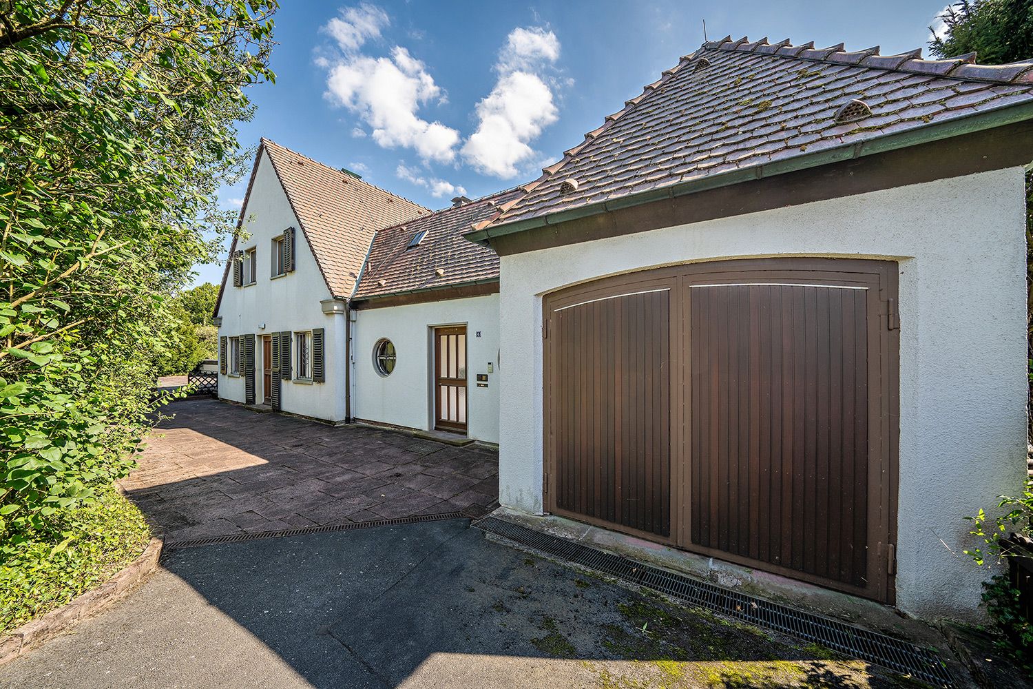 Außenansicht eines weißen Wohnhauses mit braunem Garagentor und gepflasterter Einfahrt.