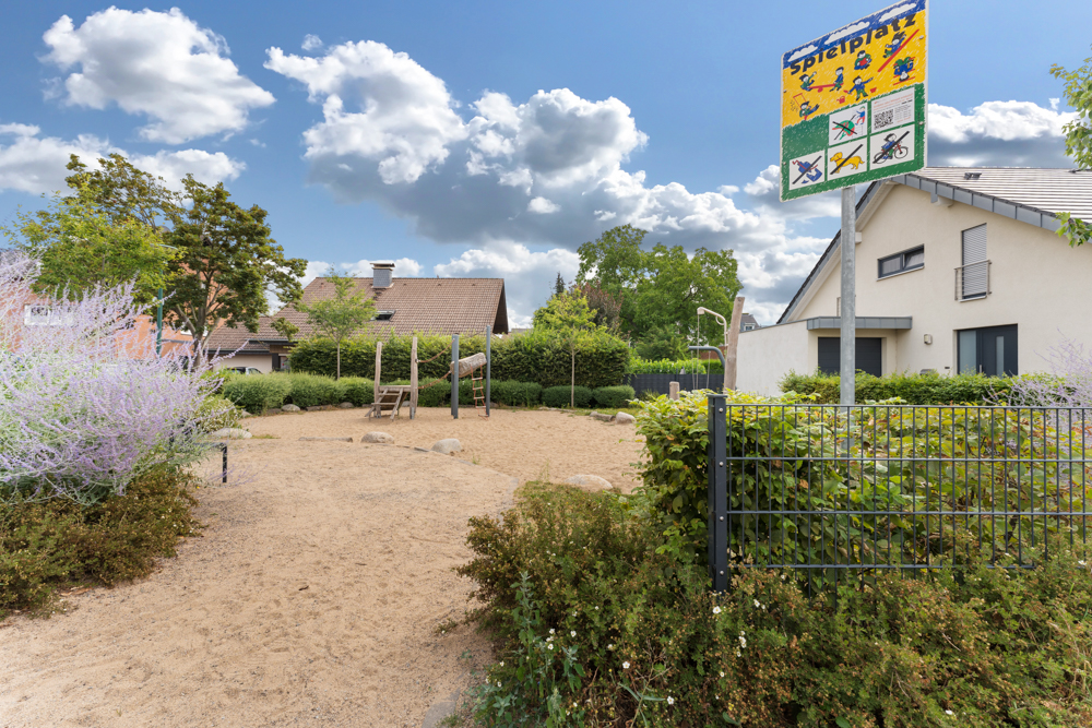 Spielplatz mit Sandfläche, Klettergerüst und Rutsche neben Wohnhäusern, im Vordergrund ein Zaun und Sträucher.