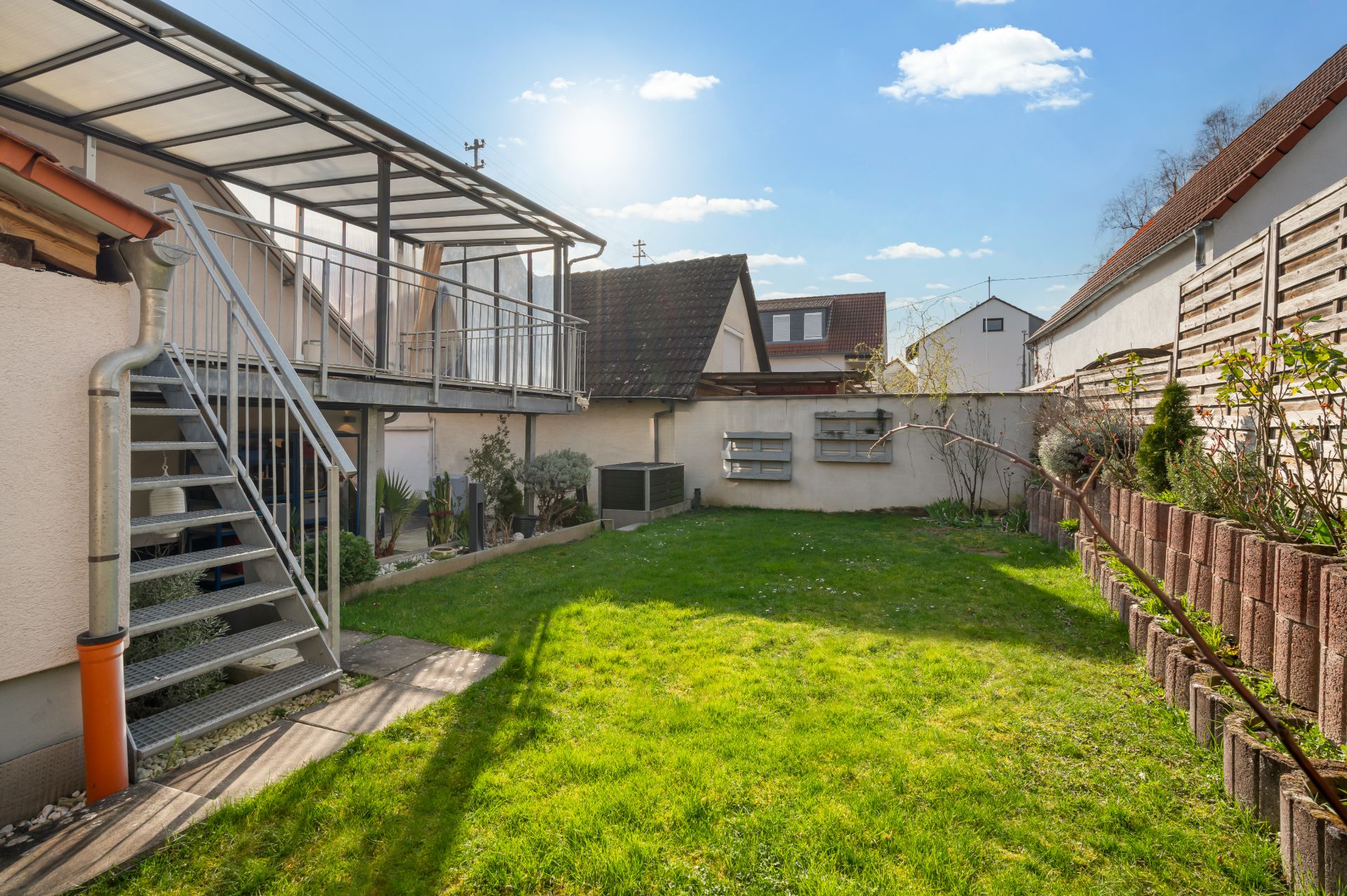 Grüner Garten mit überdachter Terrasse, Treppe und bepflanztem Randstreifen neben Wohnhäusern.