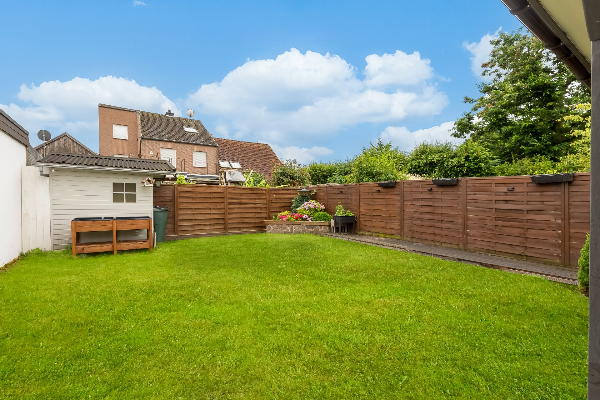 Gepflegter Garten mit Rasen, Holzzaun, Blumenbeet und kleinem weißen Schuppen.