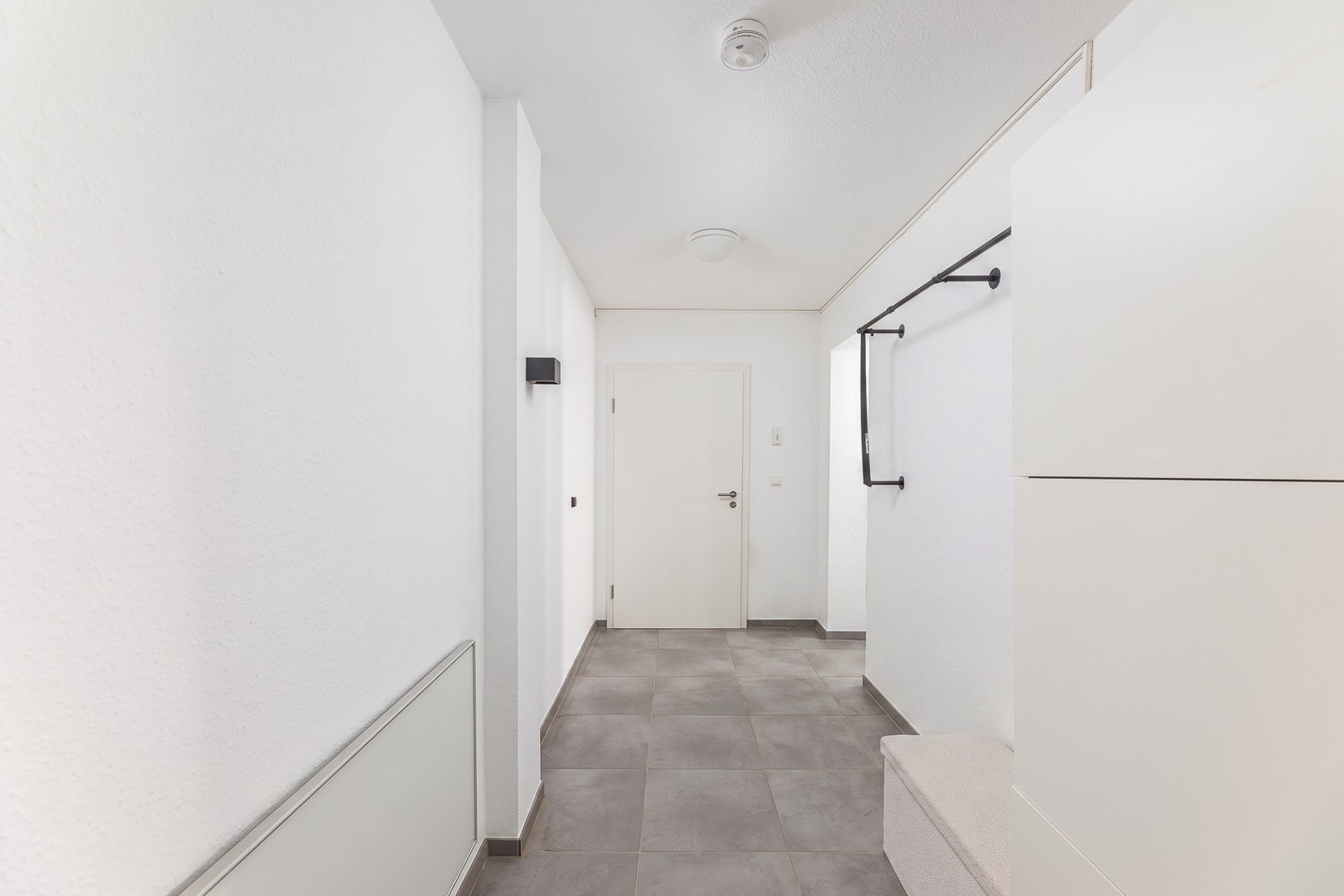 Heller Flur mit grauen Fliesen, weißer Tür und Garderobe an der Wand.