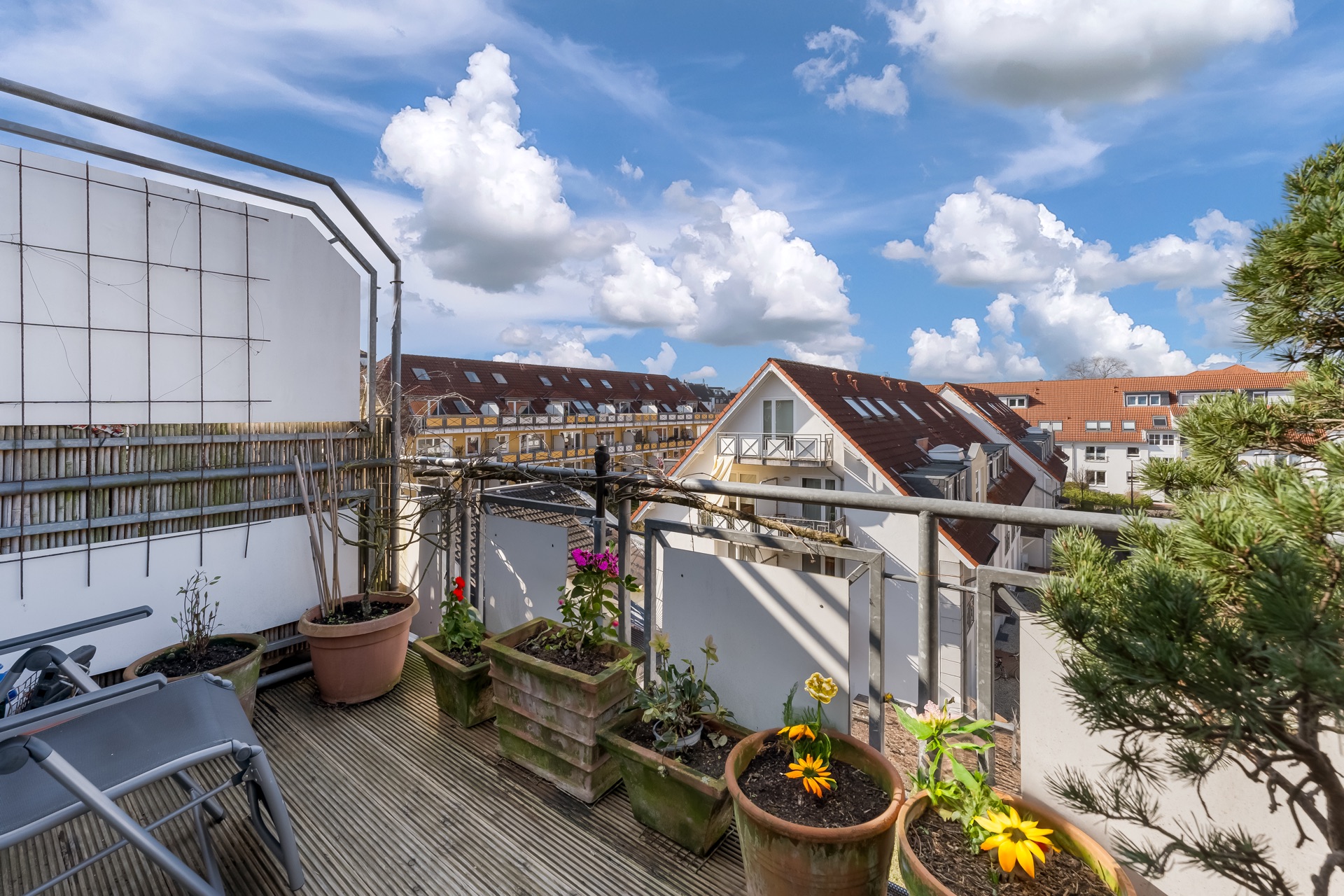 Balkon mit Blumentöpfen und Blick auf Wohnhäuser unter blauem Himmel mit Wolken.