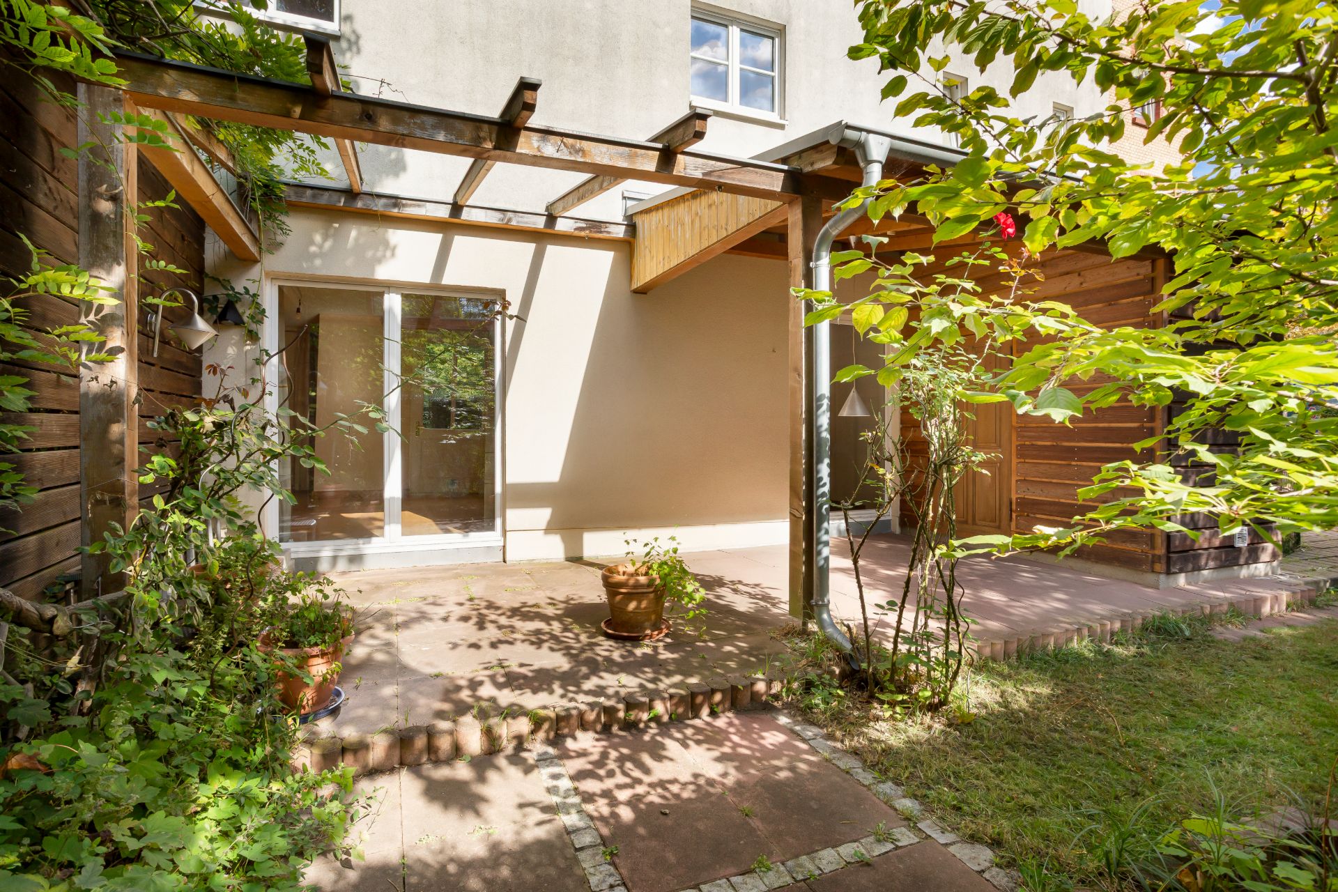 Terrasse mit Überdachung, Pflanzen in Töpfen und Zugang zum Garten an einem Wohnhaus.