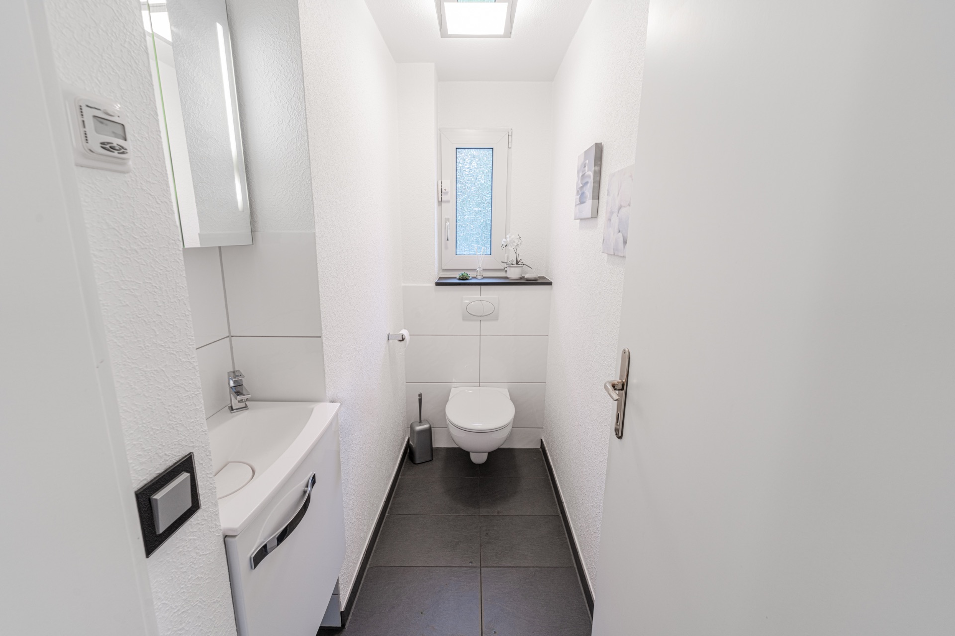 Modernes, schmales Badezimmer mit Toilette, Waschbecken und kleinem Fenster.