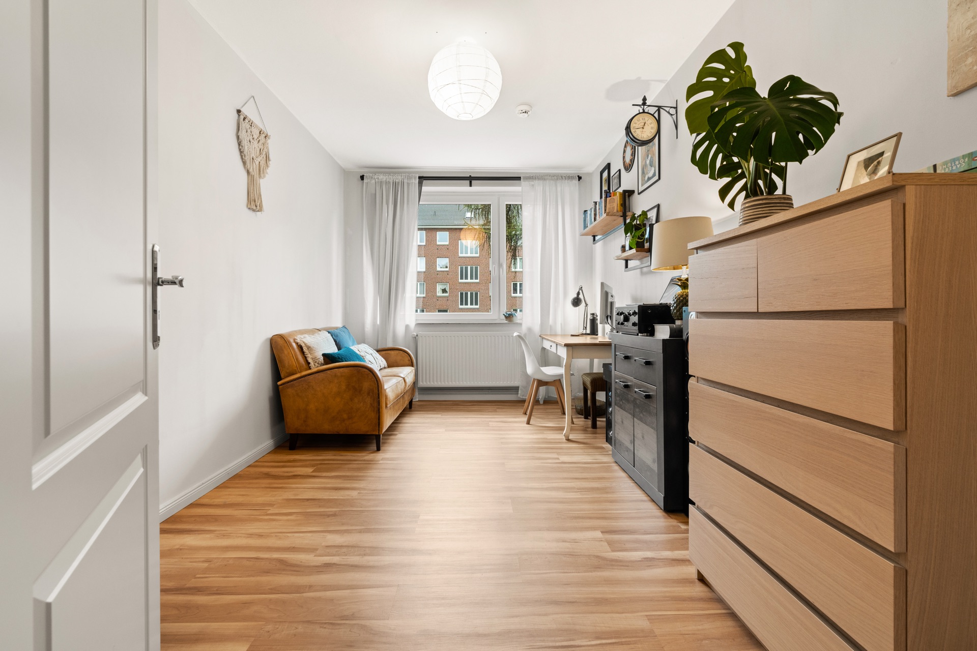 Helles Zimmer mit Holzboden, braunem Sofa, Schreibtisch, Kommode und großem Fenster.