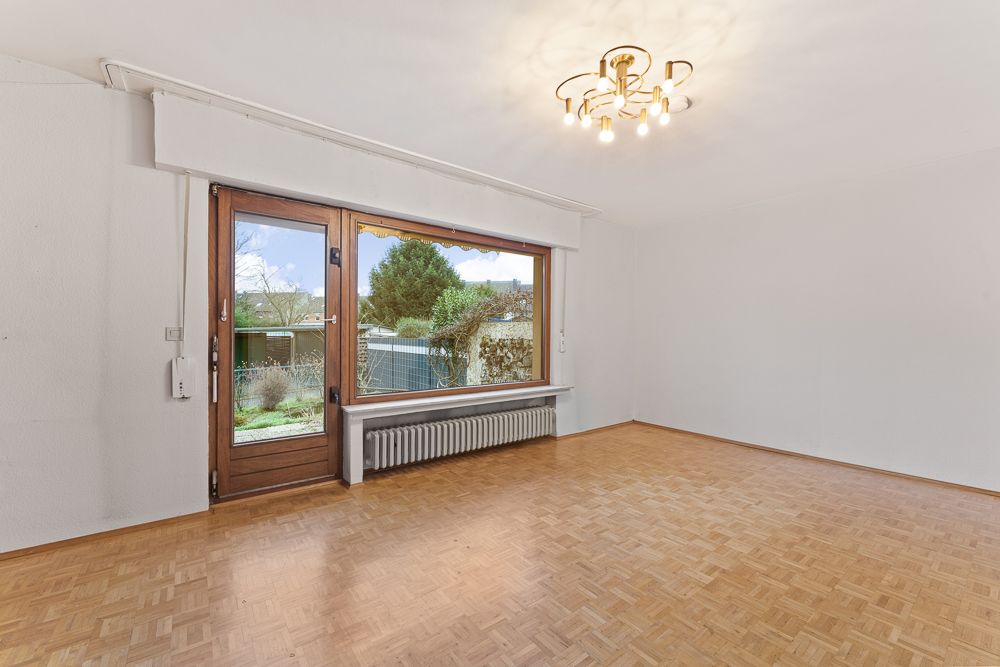 Helles, leeres Wohnzimmer mit Parkettboden, großem Fenster und Tür zum Garten.