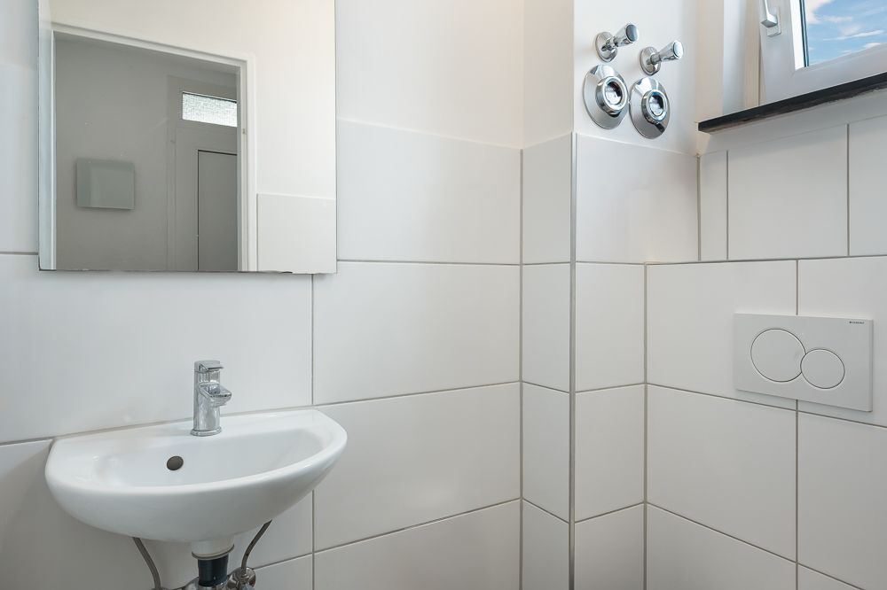 Kleines Badezimmer mit Waschbecken, Spiegel und Fenster, weiß gefliest.