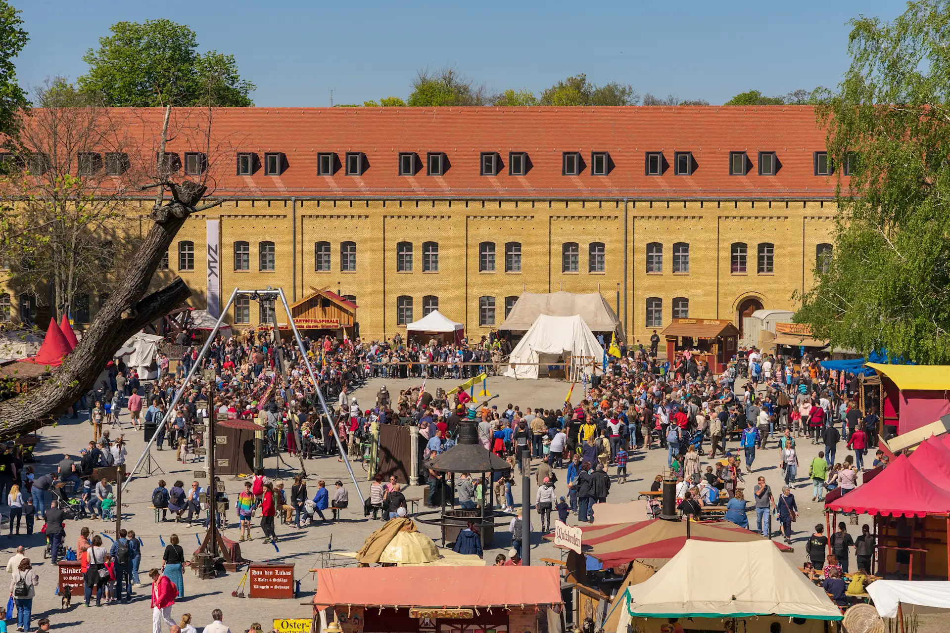 Viele Menschen auf einem belebten Marktplatz mit Ständen, Zelten und einer großen gelben Backsteinfassade im Hintergrund.