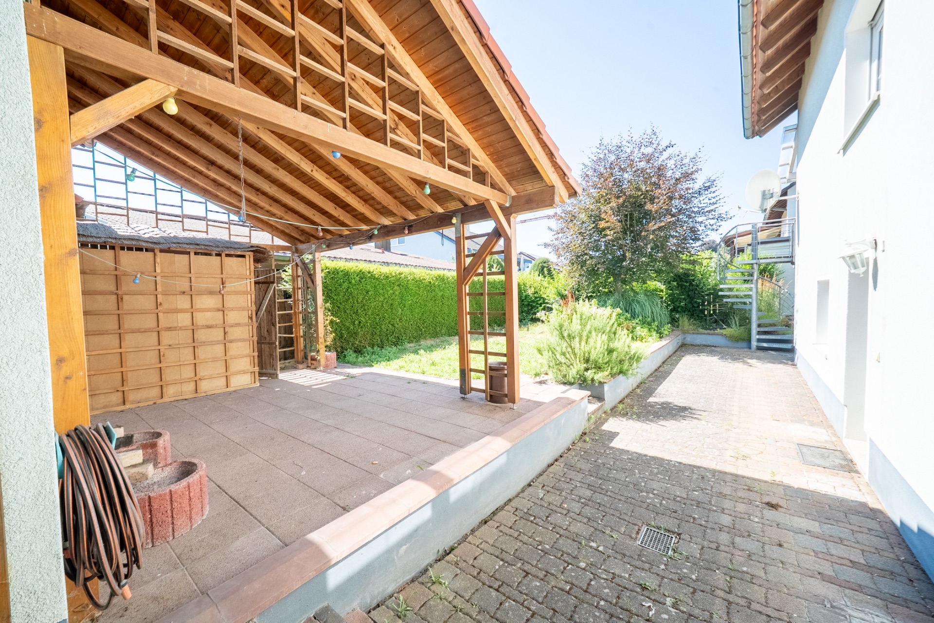 Überdachte Terrasse mit Holzdach, Gartenbereich und gepflastertem Weg neben einem Wohnhaus.