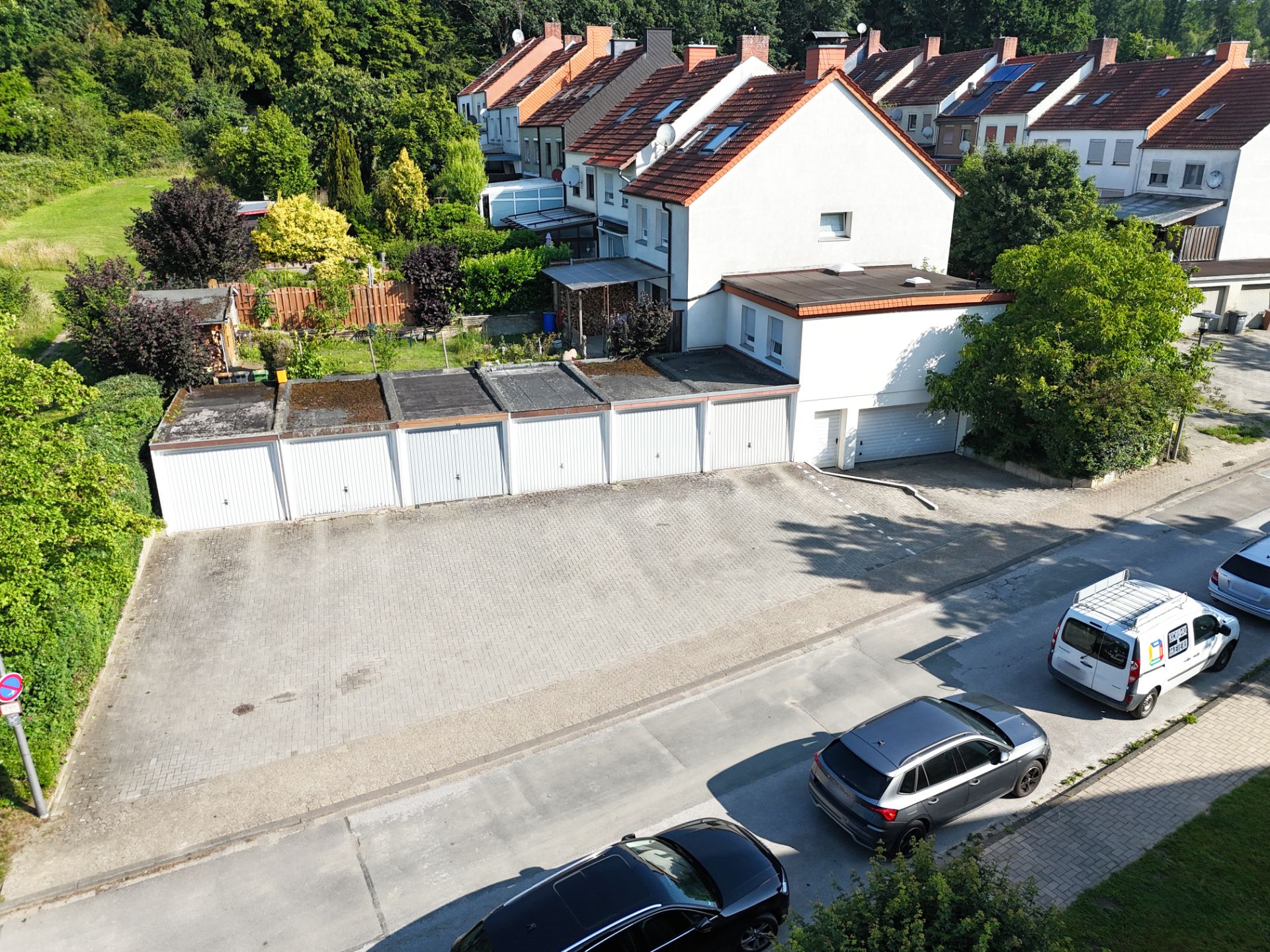 Reihenhäuser mit Garagen und geparkten Autos auf einer ruhigen Straße, im Hintergrund viel Grün.