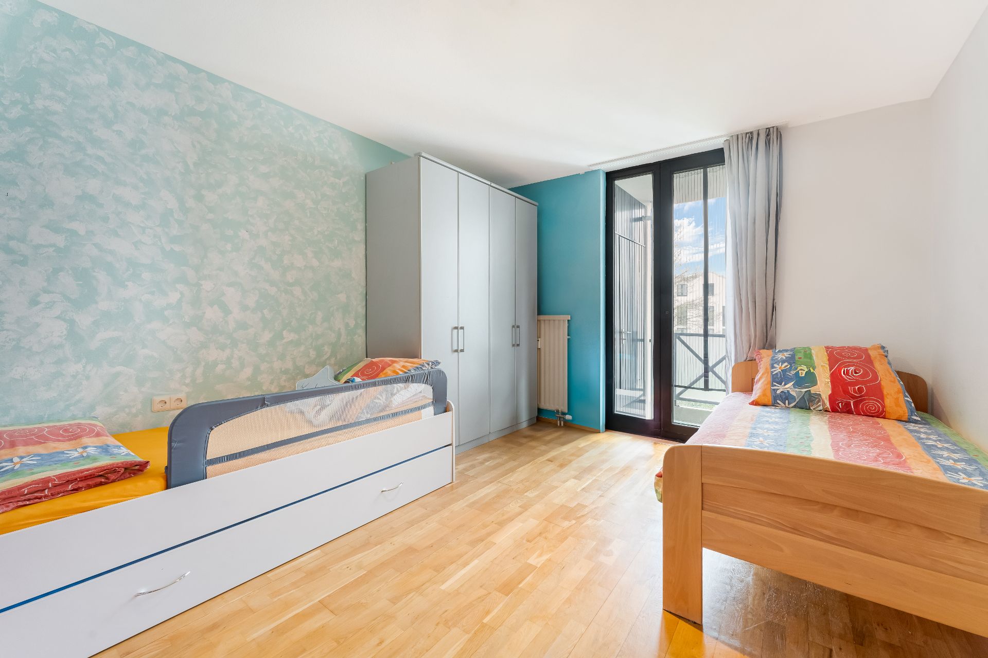 Helles Schlafzimmer mit zwei Einzelbetten, Kleiderschrank und Balkonzugang.