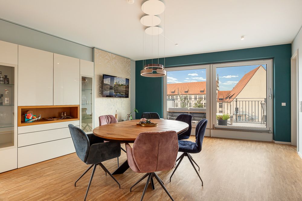 Helles Esszimmer mit rundem Holztisch, sechs Stühlen und großem Fenster mit Blick auf Dächer.