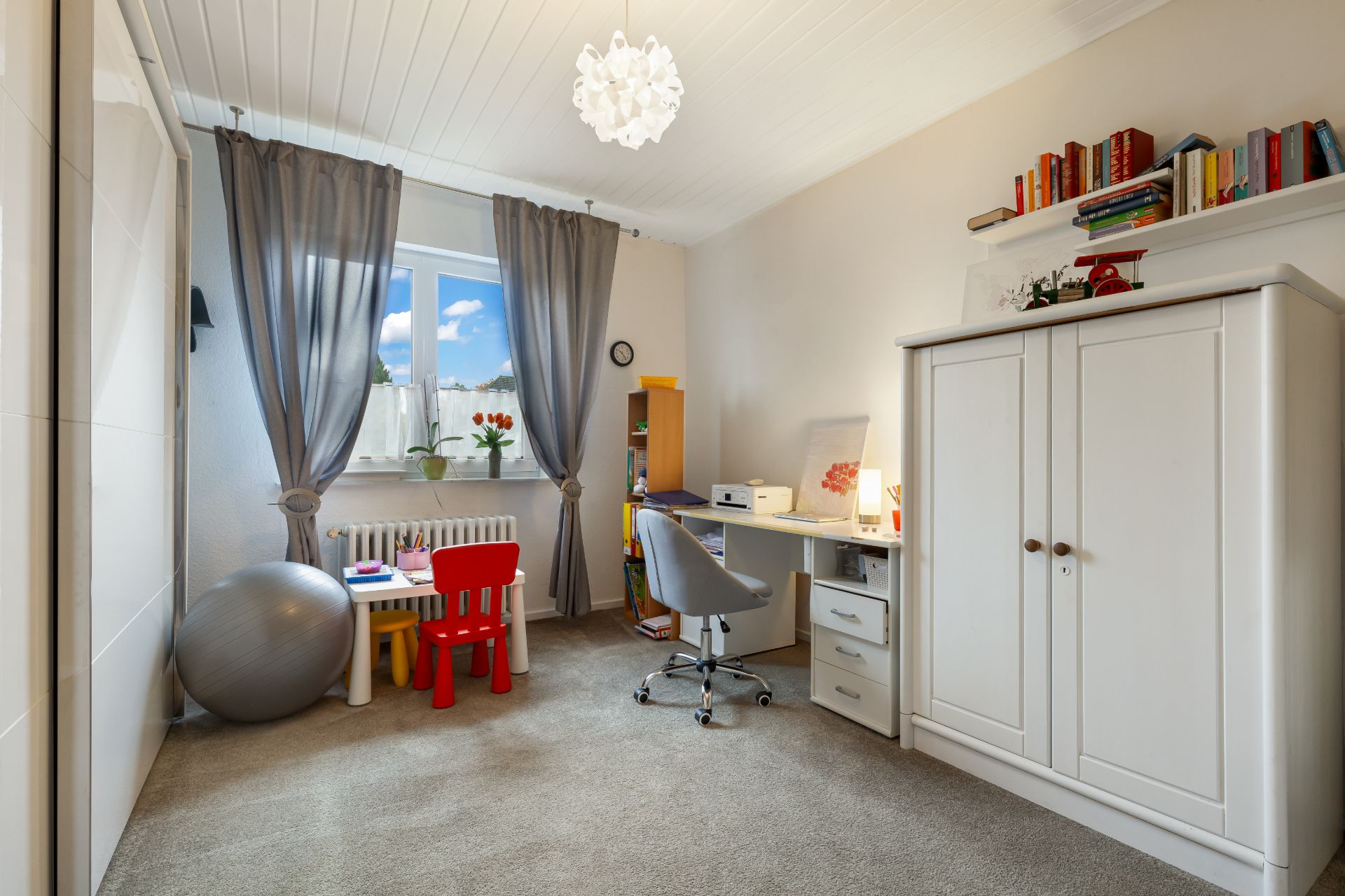 Helles Zimmer mit Schreibtisch, kleinem Kindertisch, Gymnastikball und weißem Kleiderschrank.