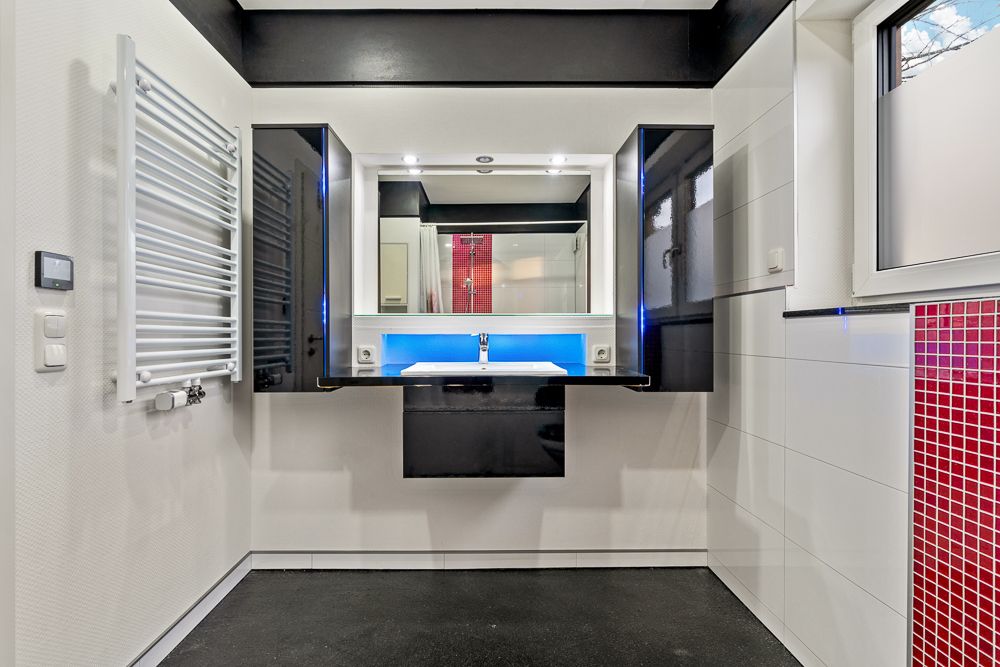 Modernes Badezimmer mit Waschbecken, großem Spiegel, schwarzen Schränken und blau beleuchtetem Regal.