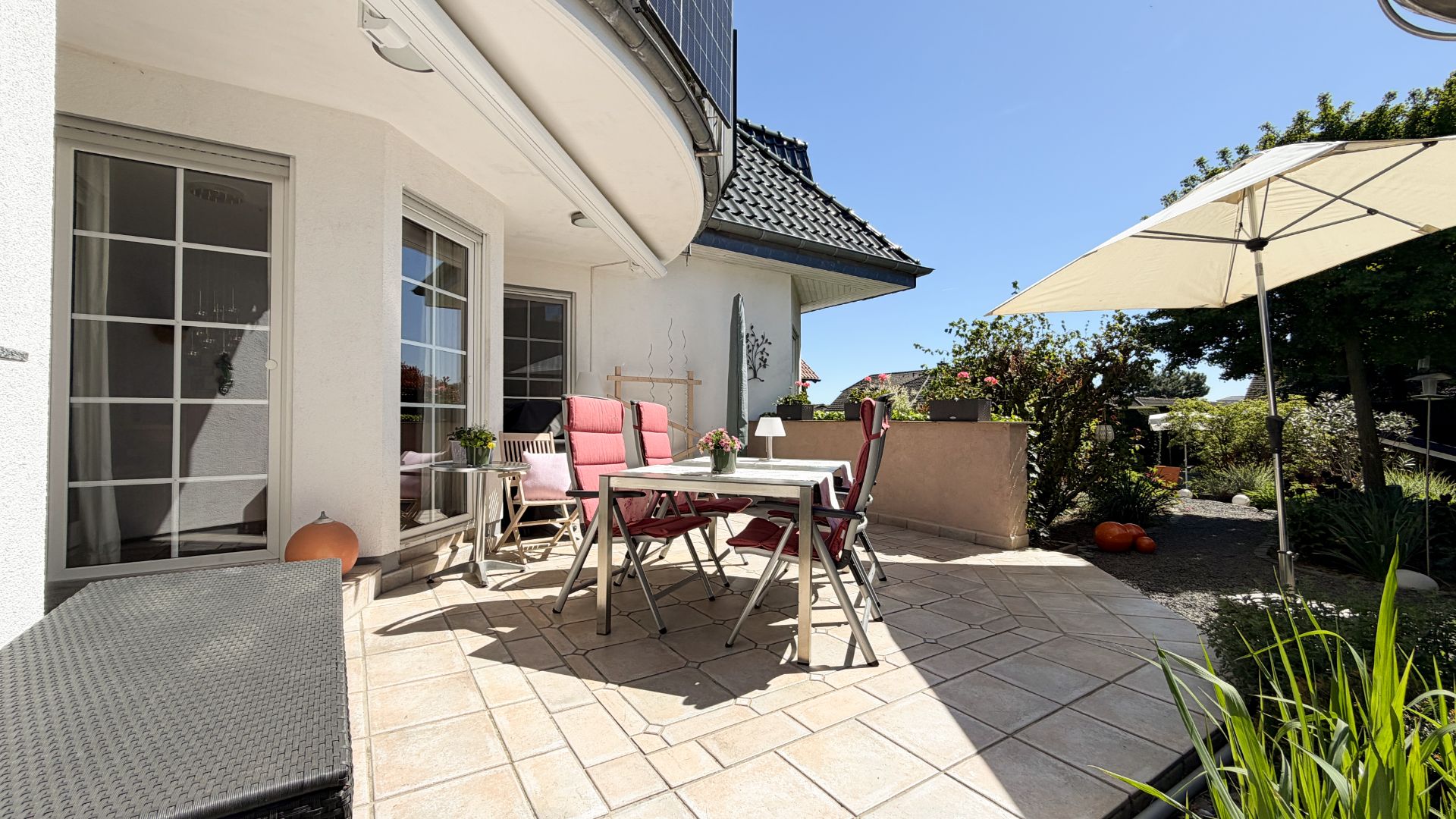 Helle Terrasse mit Tisch, Stühlen, Sonnenschirm und Garten im Hintergrund.