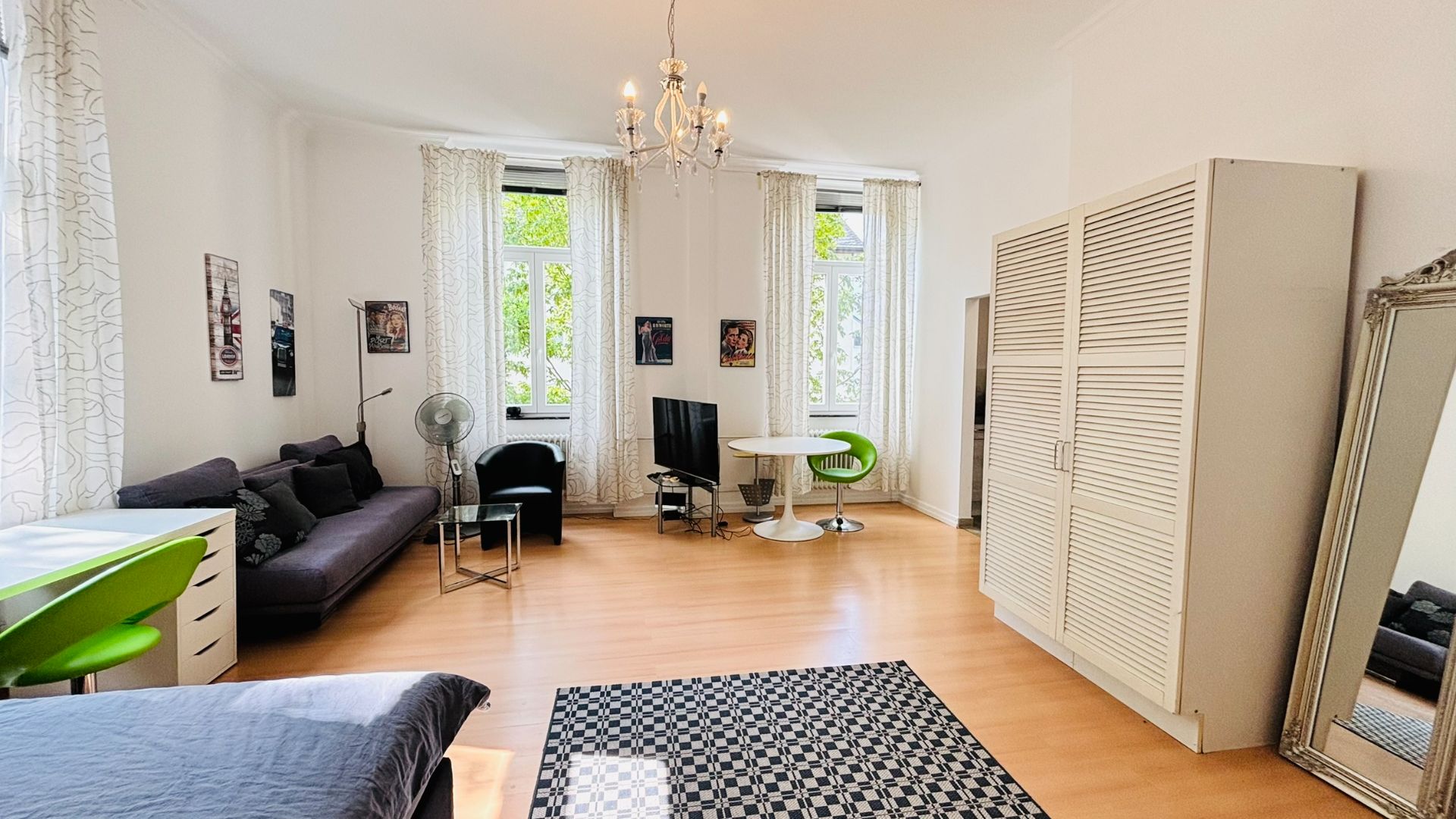 Helles Wohnzimmer mit Sofa, Schreibtisch, Tisch, Kleiderschrank und großem Spiegel.