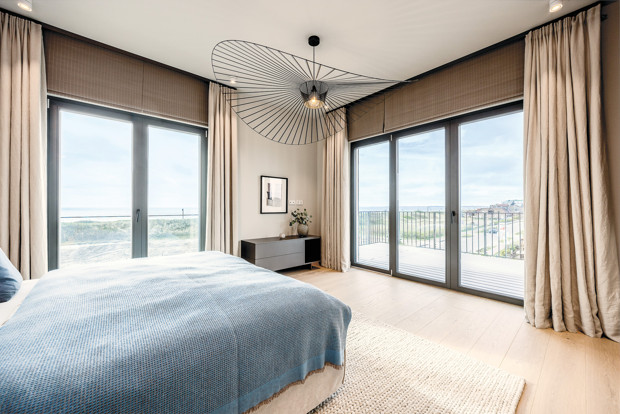 Modernes Schlafzimmer mit großem Bett, Holzboden und bodentiefen Fenstern mit Blick auf Balkon und Natur.