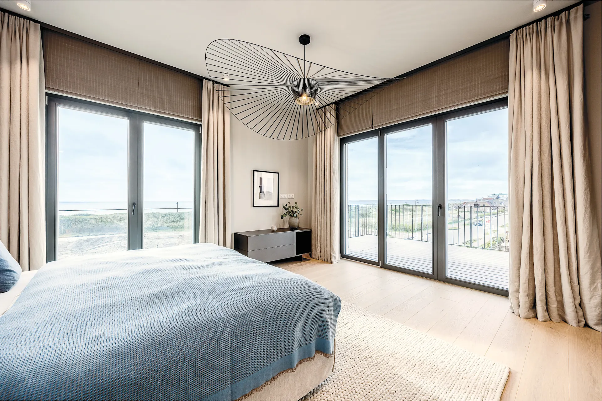 Modernes Schlafzimmer mit großem Bett, Holzboden und bodentiefen Fenstern mit Blick auf Balkon und Natur.