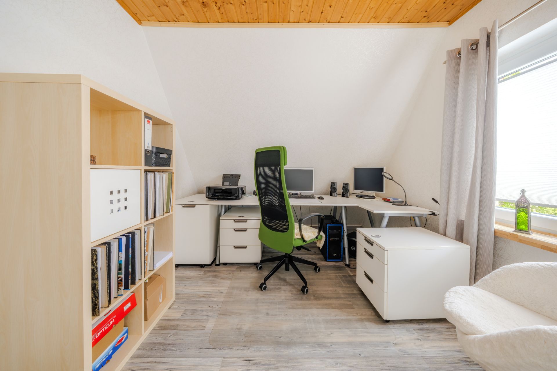 Helles Arbeitszimmer mit Schreibtisch, zwei Monitoren, grünem Stuhl und Bücherregal.