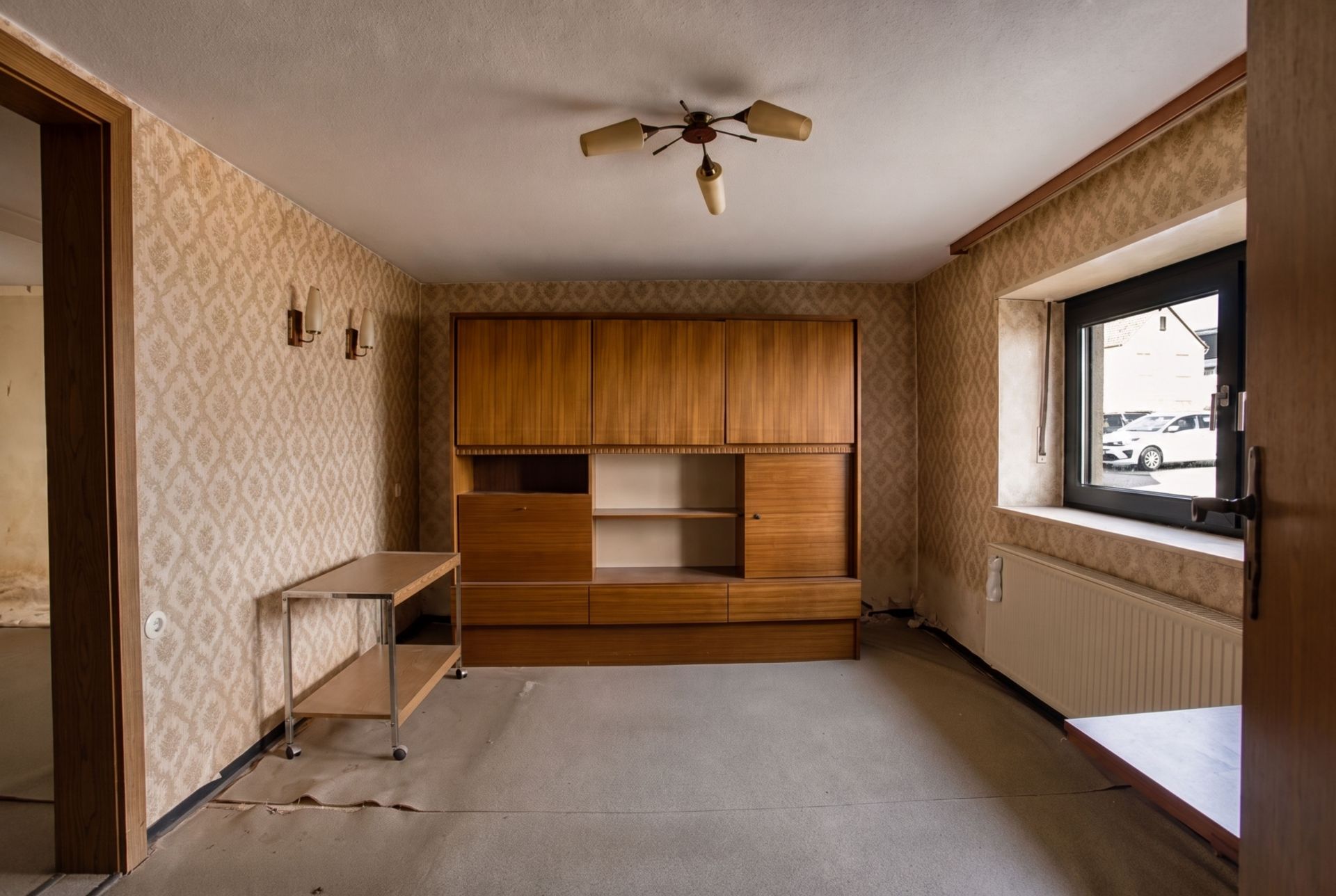 Wohnzimmer mit Holzschrank, Tisch, Fenster und Tapeten mit Muster.