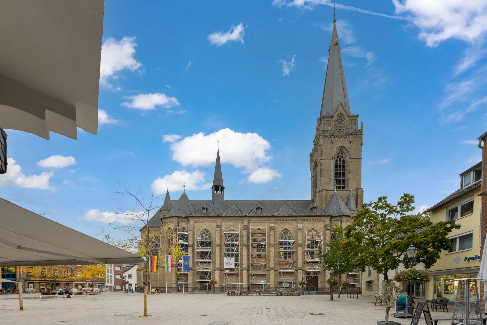 Große Kirche mit Gerüst an der Fassade, auf einem offenen Platz mit einigen Bäumen und Gebäuden.