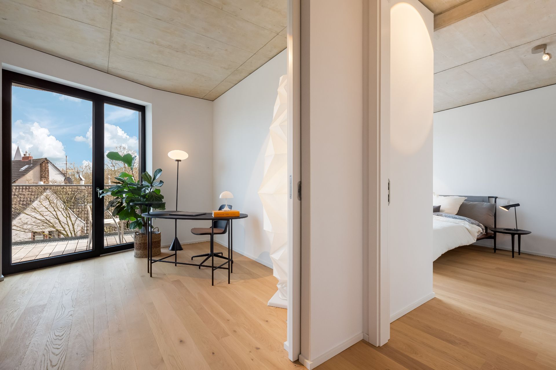 Heller Raum mit Schreibtisch und Pflanze, daneben ein Schlafzimmer mit Bett auf Holzboden.