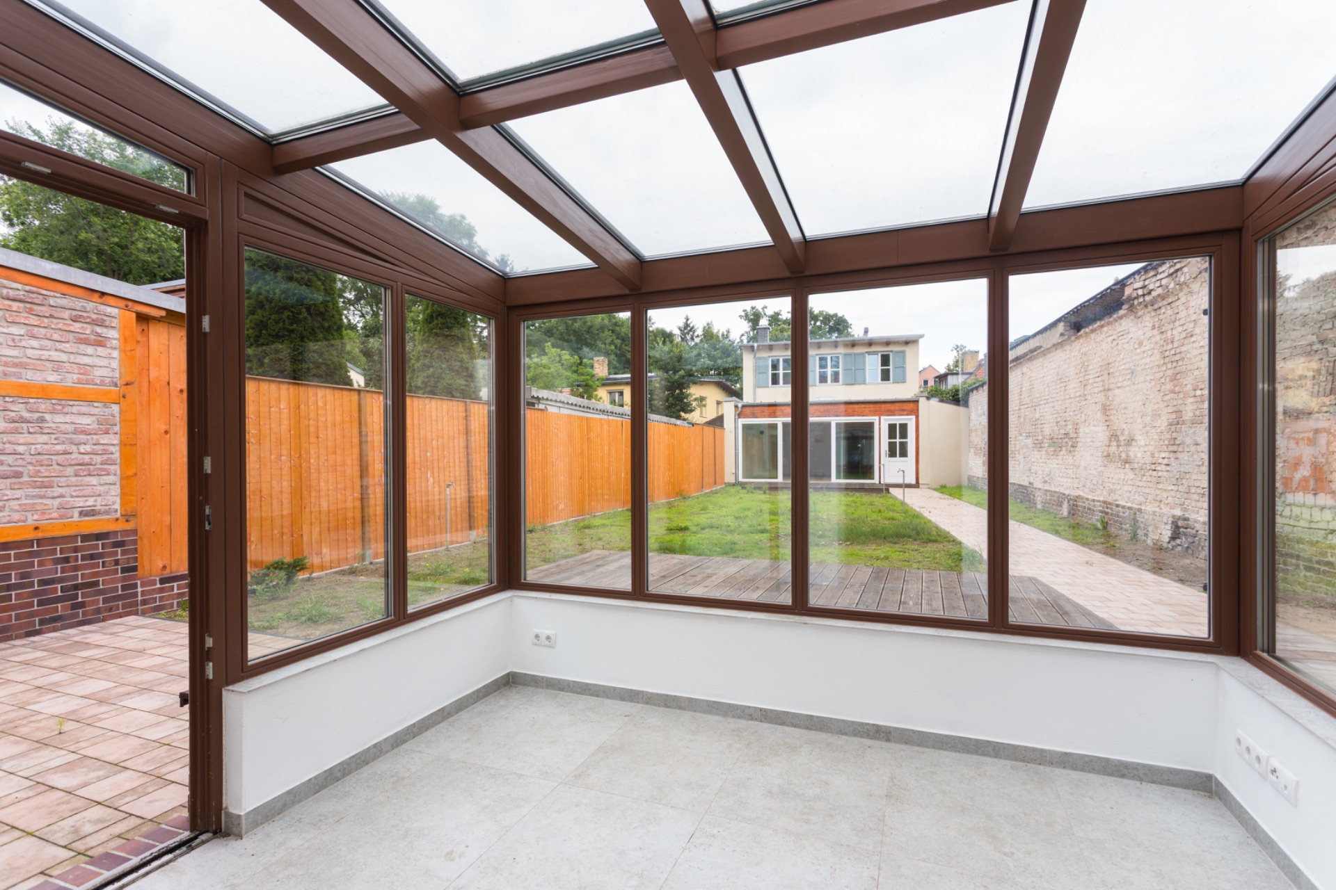 Wintergarten mit Glaswänden und Blick auf einen Garten mit Holzzaun und Haus im Hintergrund.