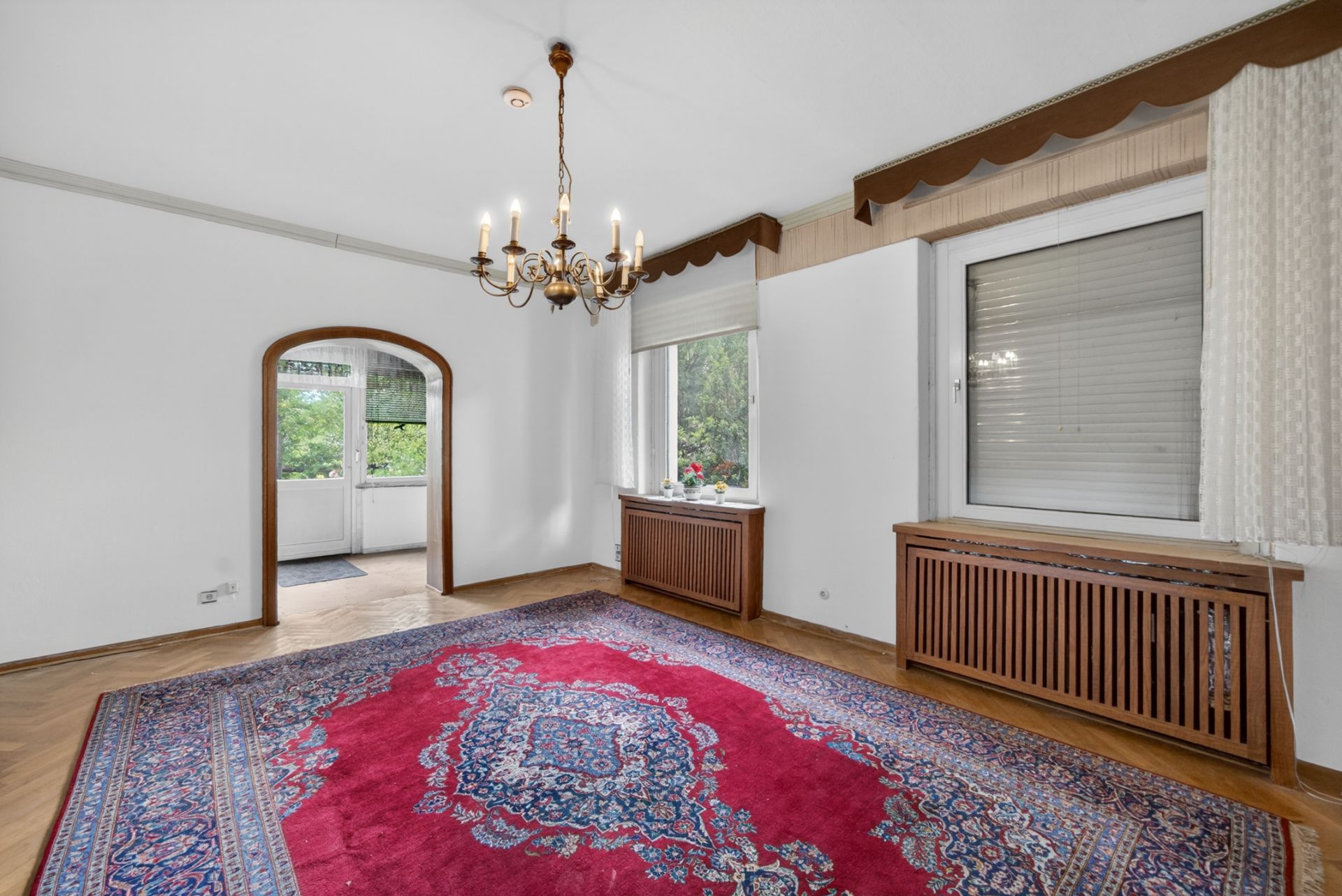 Wohnzimmer mit großem roten Teppich, Kronleuchter, zwei Fenstern und Durchgang zu weiterem Raum.