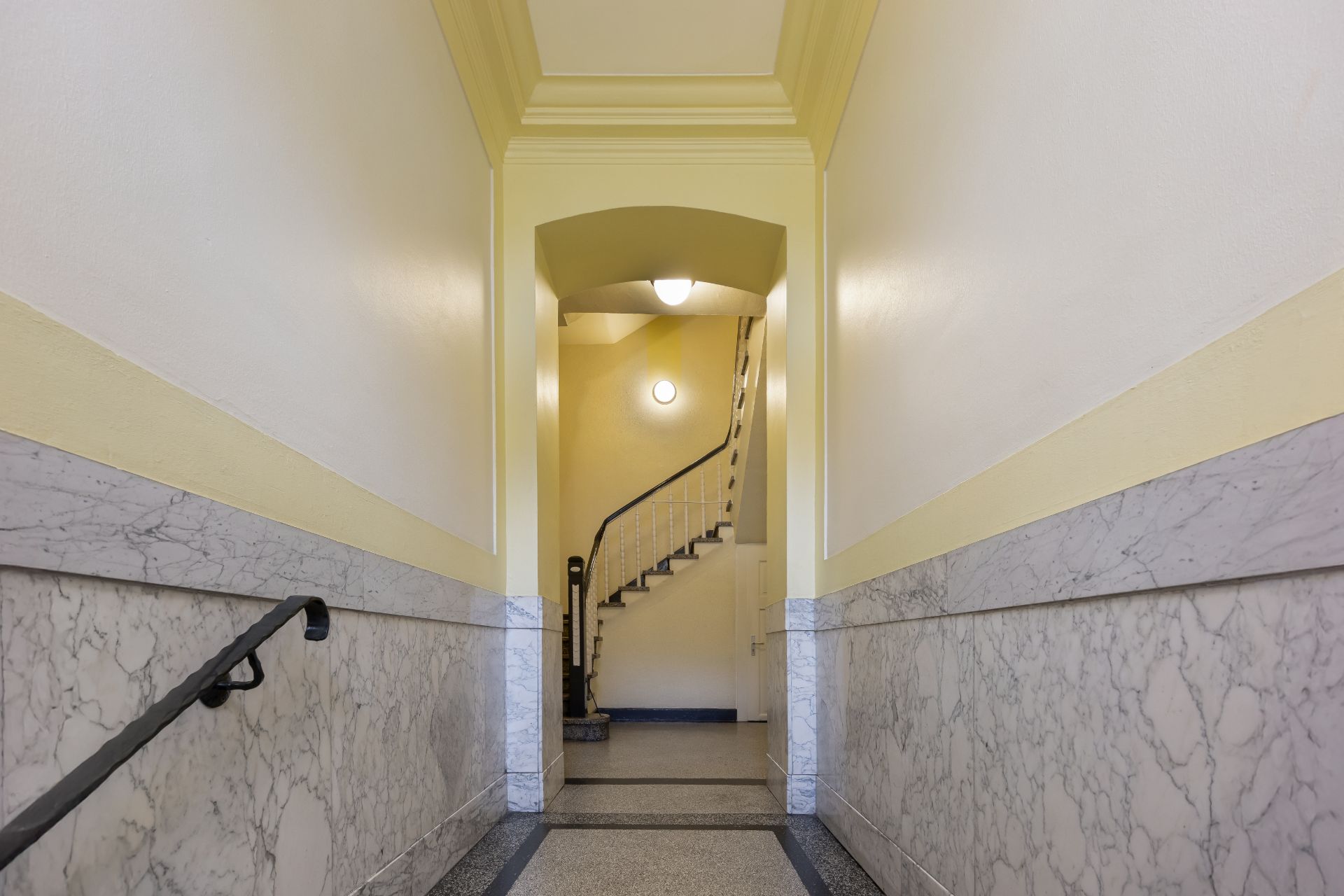 Heller Hausflur mit Marmordekor und Treppe, Wandlampen beleuchten den Bereich.