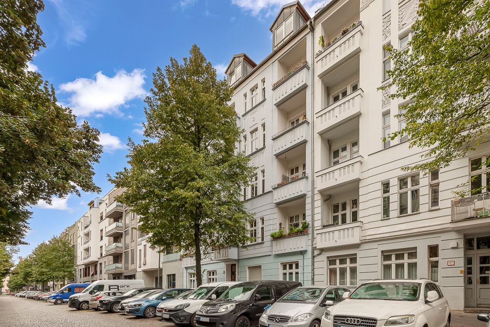 Mehrstöckiges Wohnhaus mit Balkonen und Bäumen entlang einer Straße, Autos parken am Straßenrand.