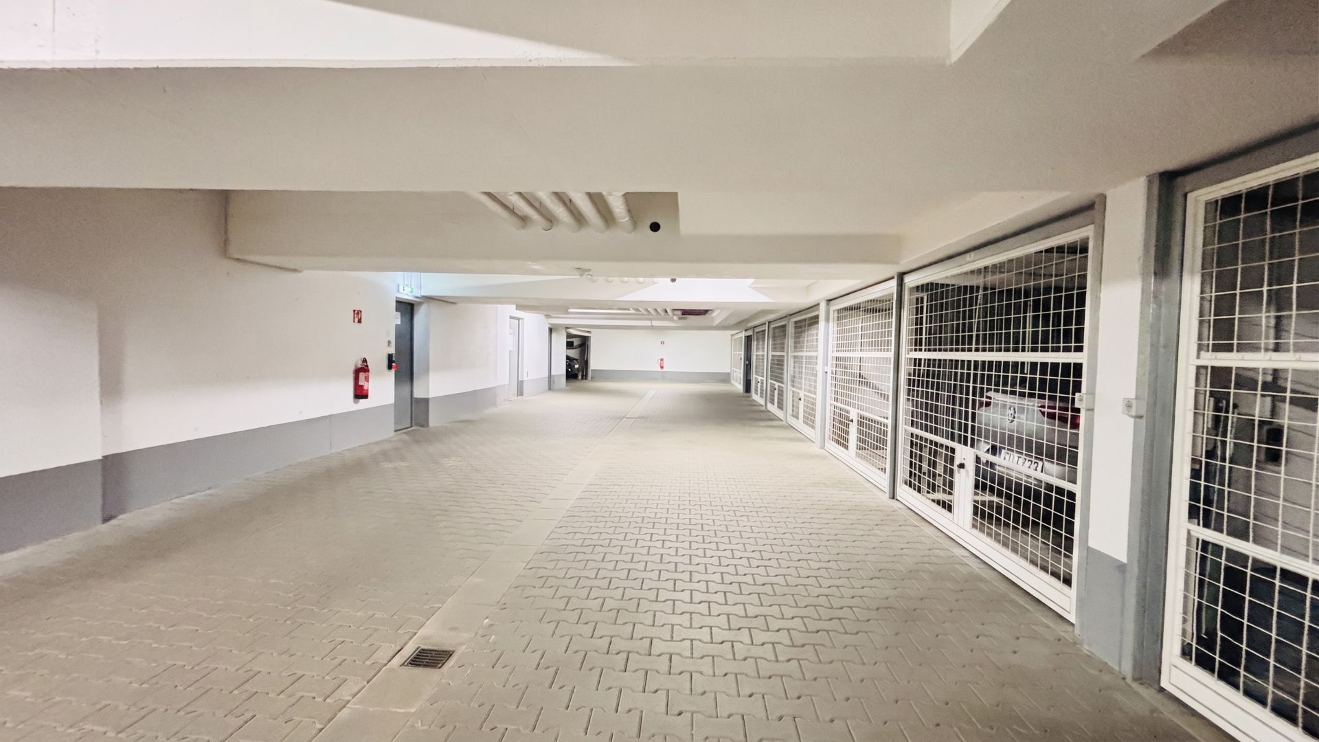 Helle Tiefgarage mit Gittertüren zu einzelnen Stellplätzen und Feuerlöschern an der Wand.