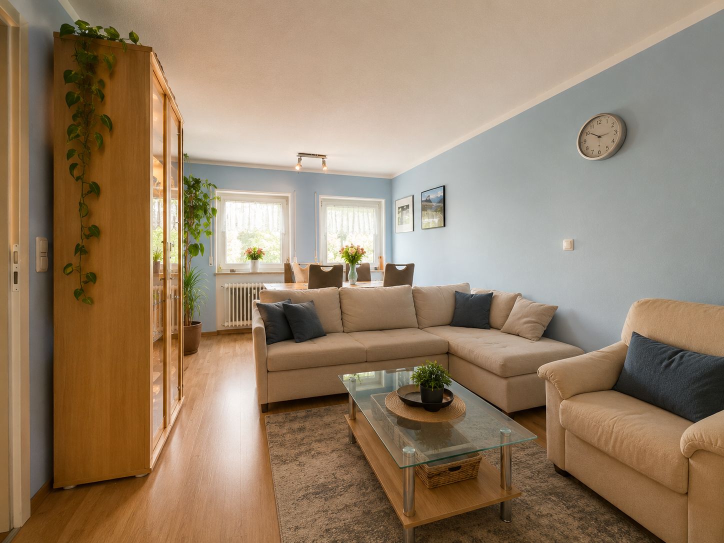 Helles Wohnzimmer mit Ecksofa, Sessel, Glastisch und Esstisch vor zwei Fenstern.