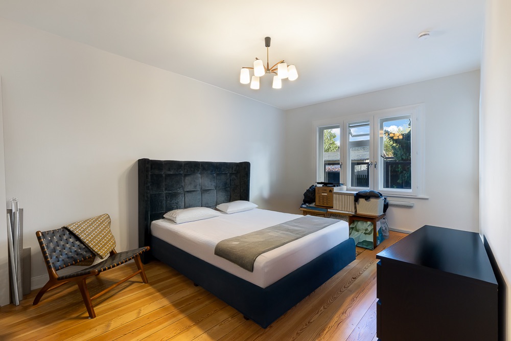 Schlafzimmer mit Doppelbett, Stuhl, Kommode und drei Fenstern, Holzboden.