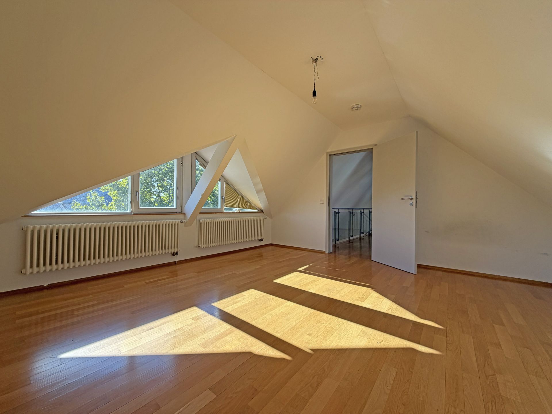 Leeres Dachzimmer mit Parkettboden, Fenstern und offener Tür, Sonnenlicht fällt auf den Boden.
