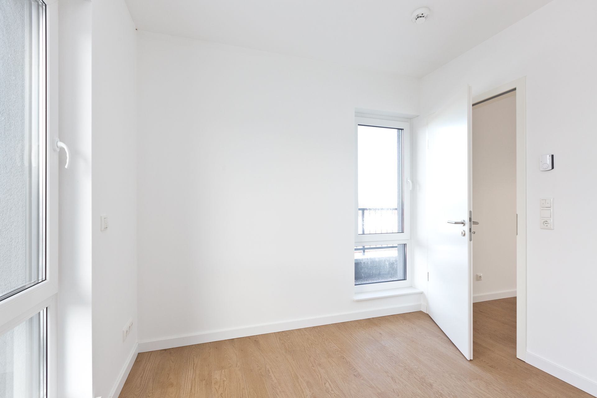 Heller Raum mit weißer Wand, Holzboden, Fenster und geöffneter Tür.