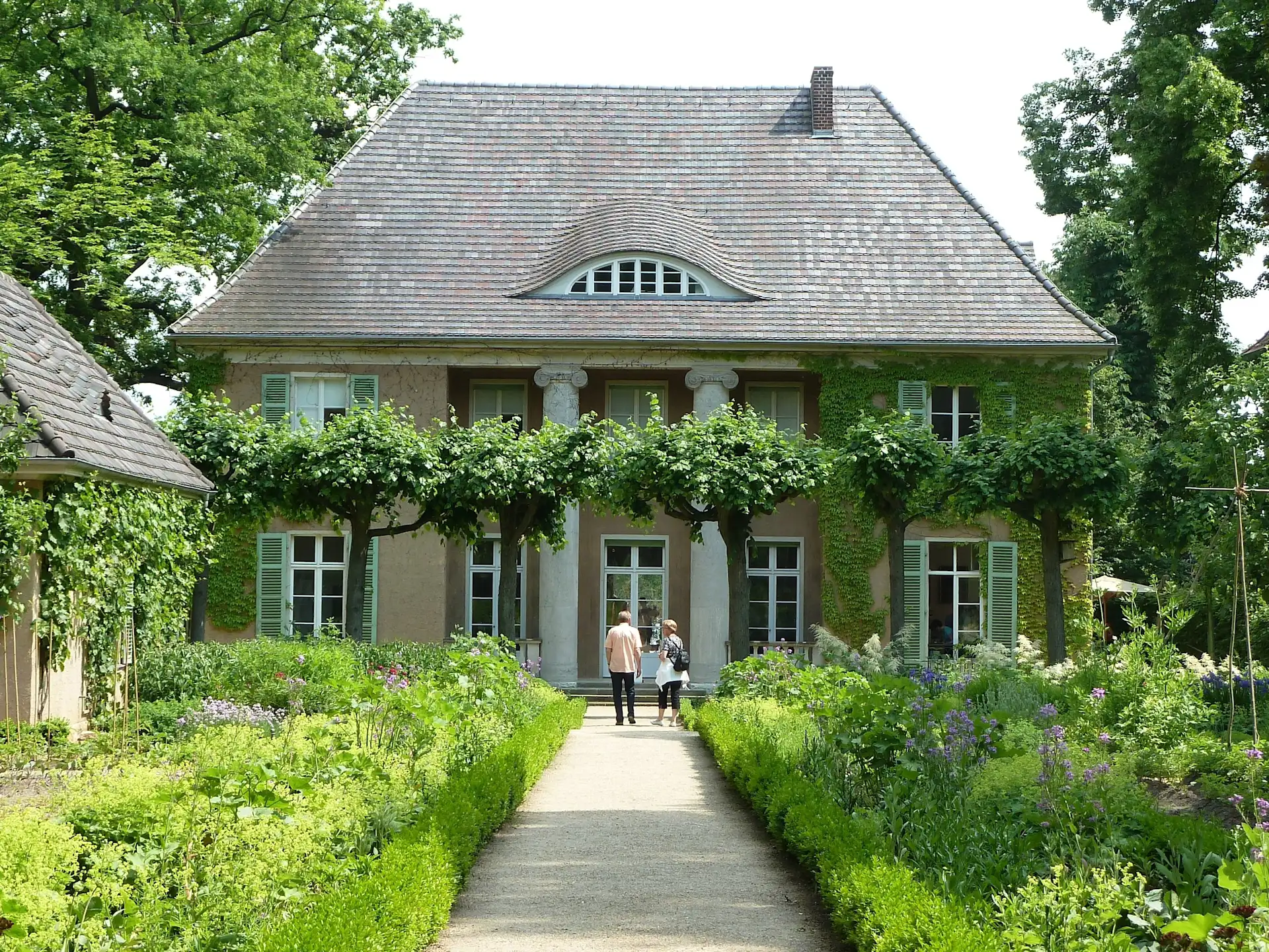 Großes Haus mit grün bewachsenen Wänden, Gartenweg und zwei Personen im Vordergrund.