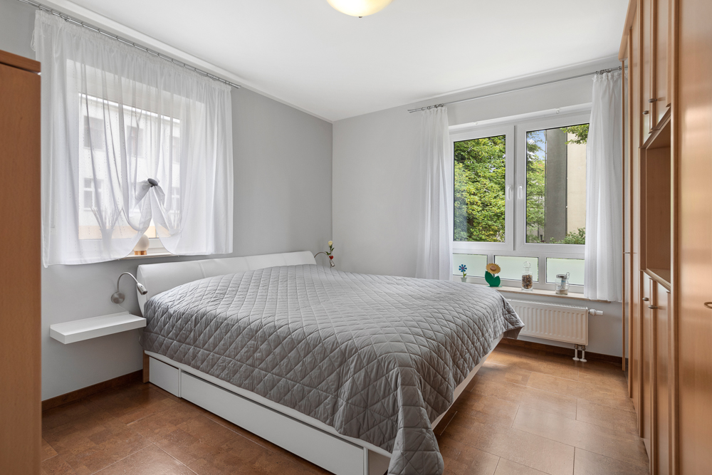 Helles Schlafzimmer mit großem Bett, Holzschränken und zwei Fenstern mit weißen Vorhängen.