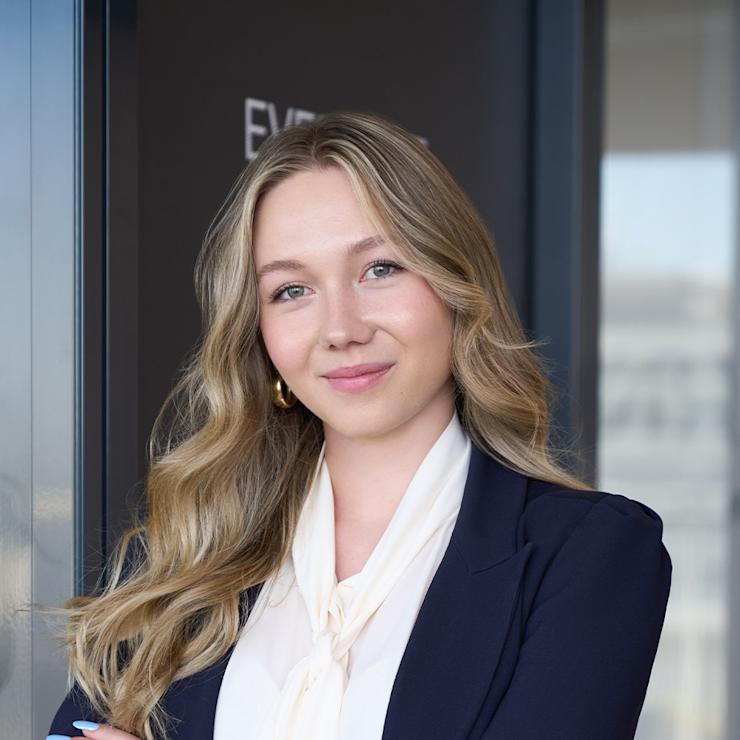 Junge Frau mit langen blonden Haaren und dunklem Blazer steht mit verschränkten Armen.