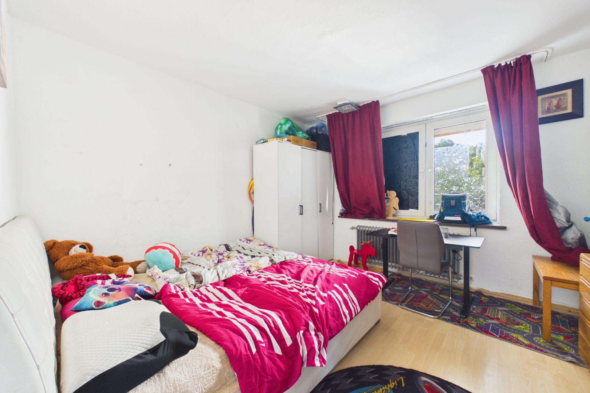 Kinderzimmer mit Bett, bunten Decken, Kuscheltieren, Schrank und Schreibtisch am Fenster mit roten Vorhängen.