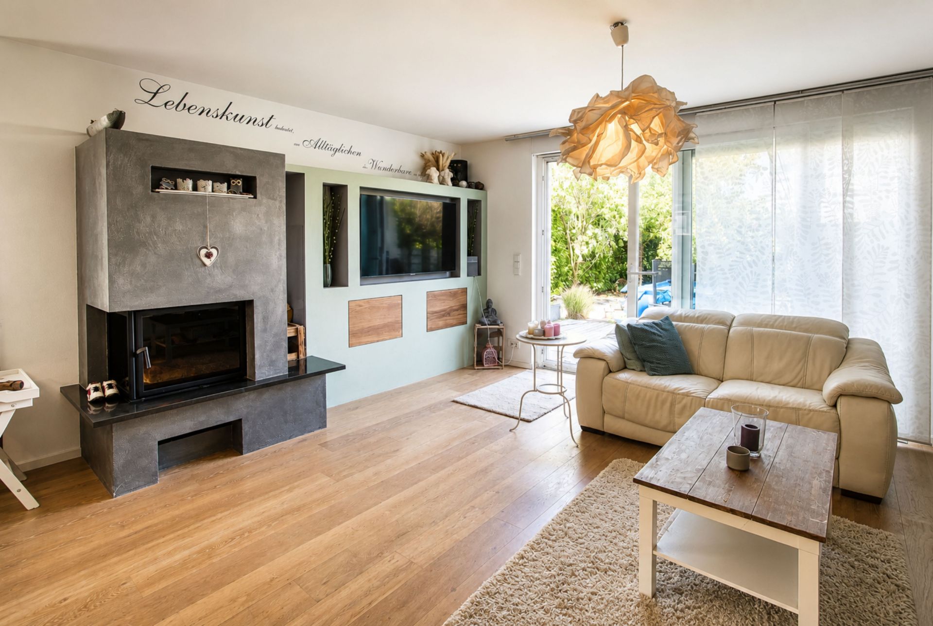 Helles Wohnzimmer mit Kamin, Sofa, Couchtisch und großem Fenster mit Blick ins Grüne.