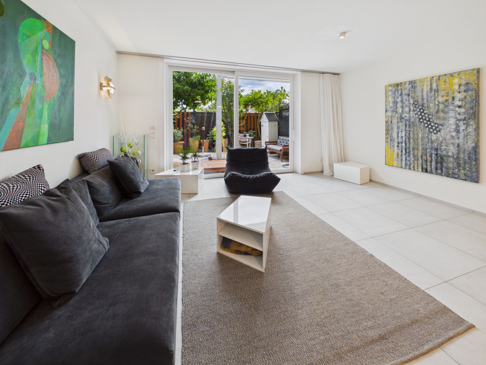 Helles Wohnzimmer mit grauem Sofa, Teppich, modernen Möbeln und Zugang zu einer Terrasse mit Garten.