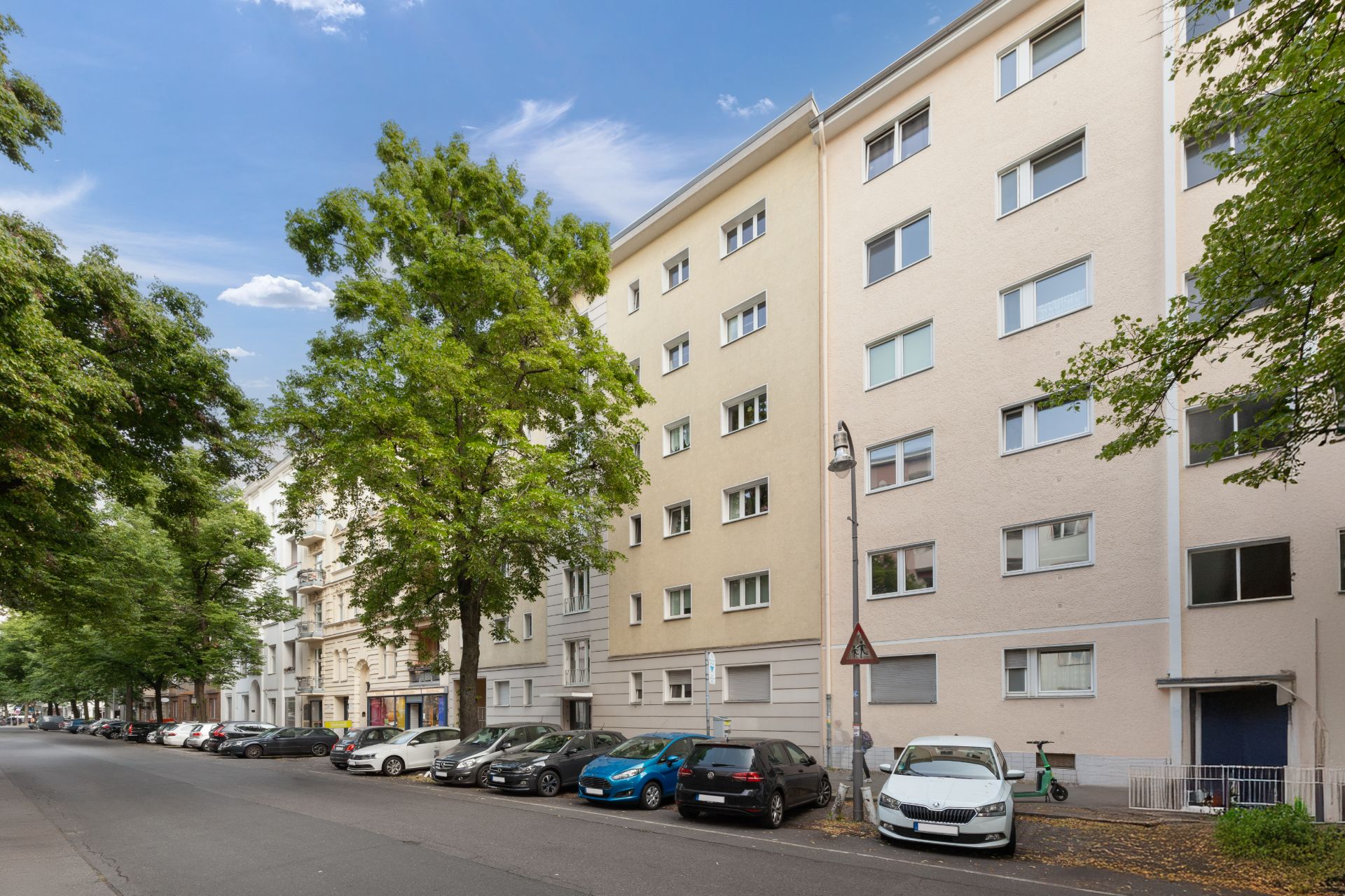 Mehrstöckiges Wohnhaus mit geparkten Autos und Bäumen an einer ruhigen Straße.