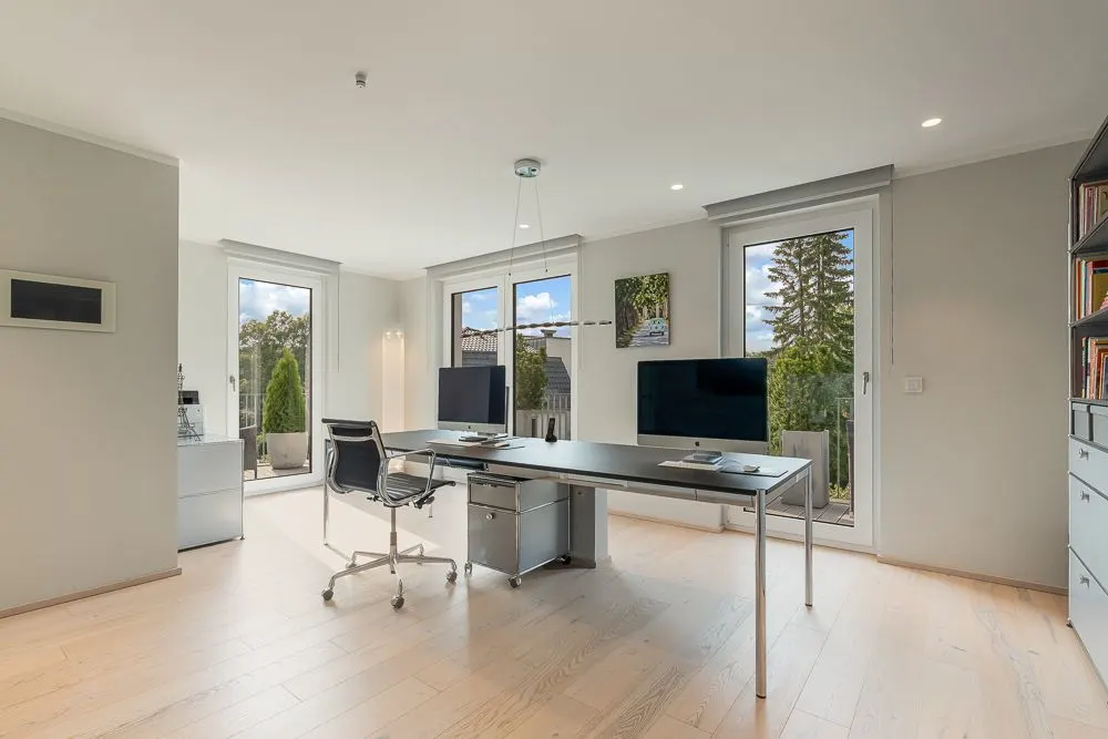 Helles Arbeitszimmer mit großem Schreibtisch, Stuhl und zwei Monitoren vor Fenstern.