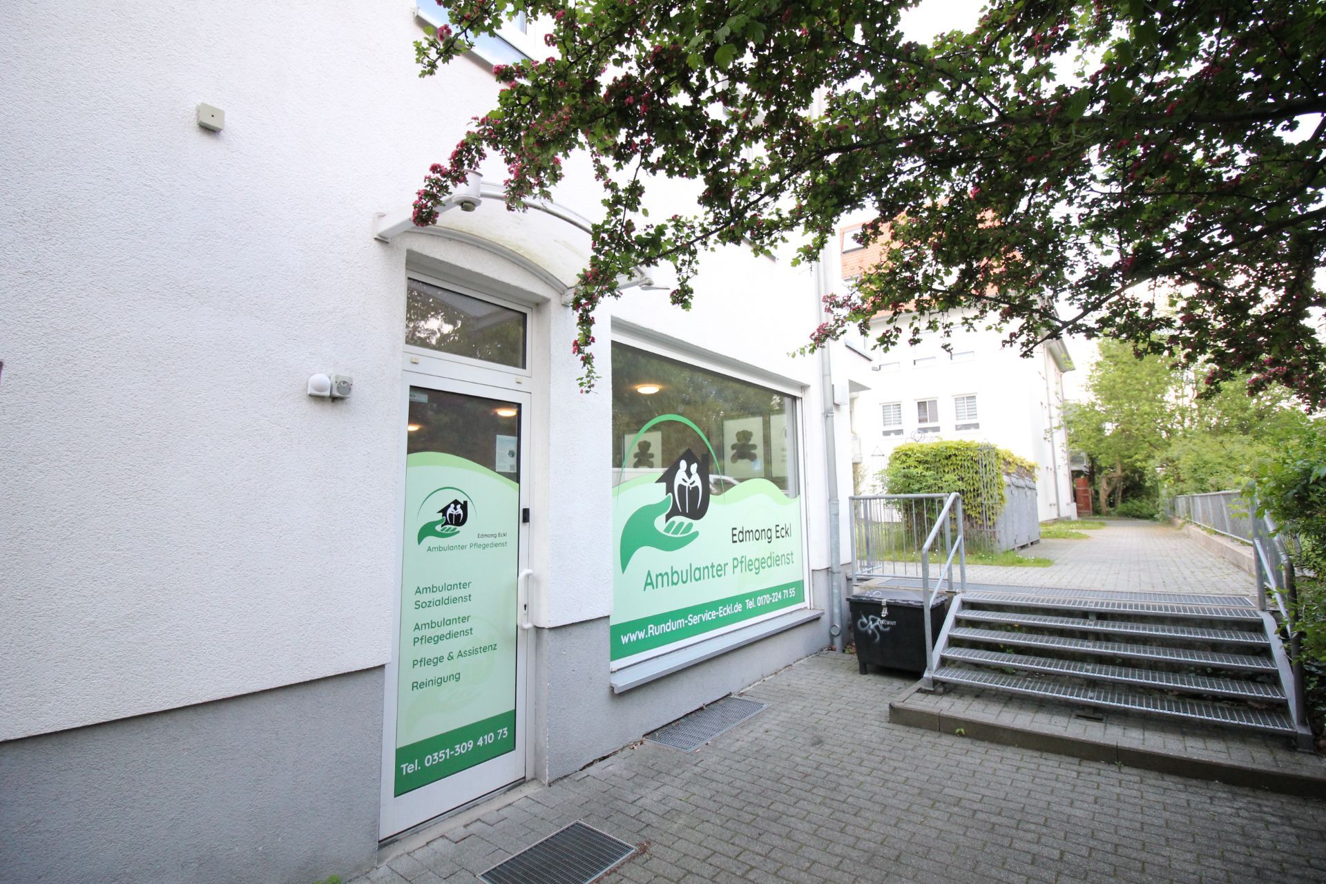 Eingang zu einem ambulanten Pflegedienst mit Beschilderung an Fenster und Tür, Gehweg und Bäume im Vordergrund.