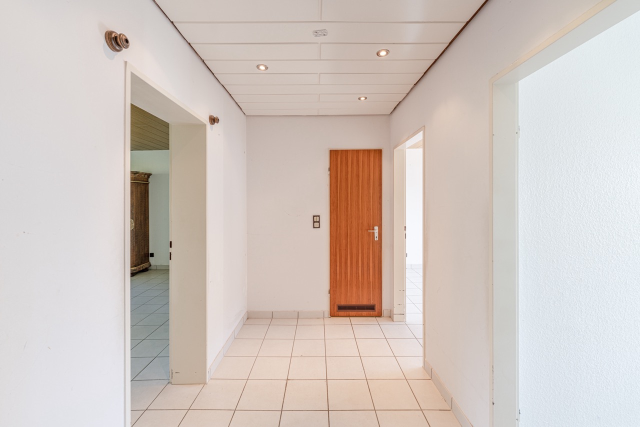 Heller Flur mit weißen Wänden, weißen Fliesen und Holztür in einer Wohnung.