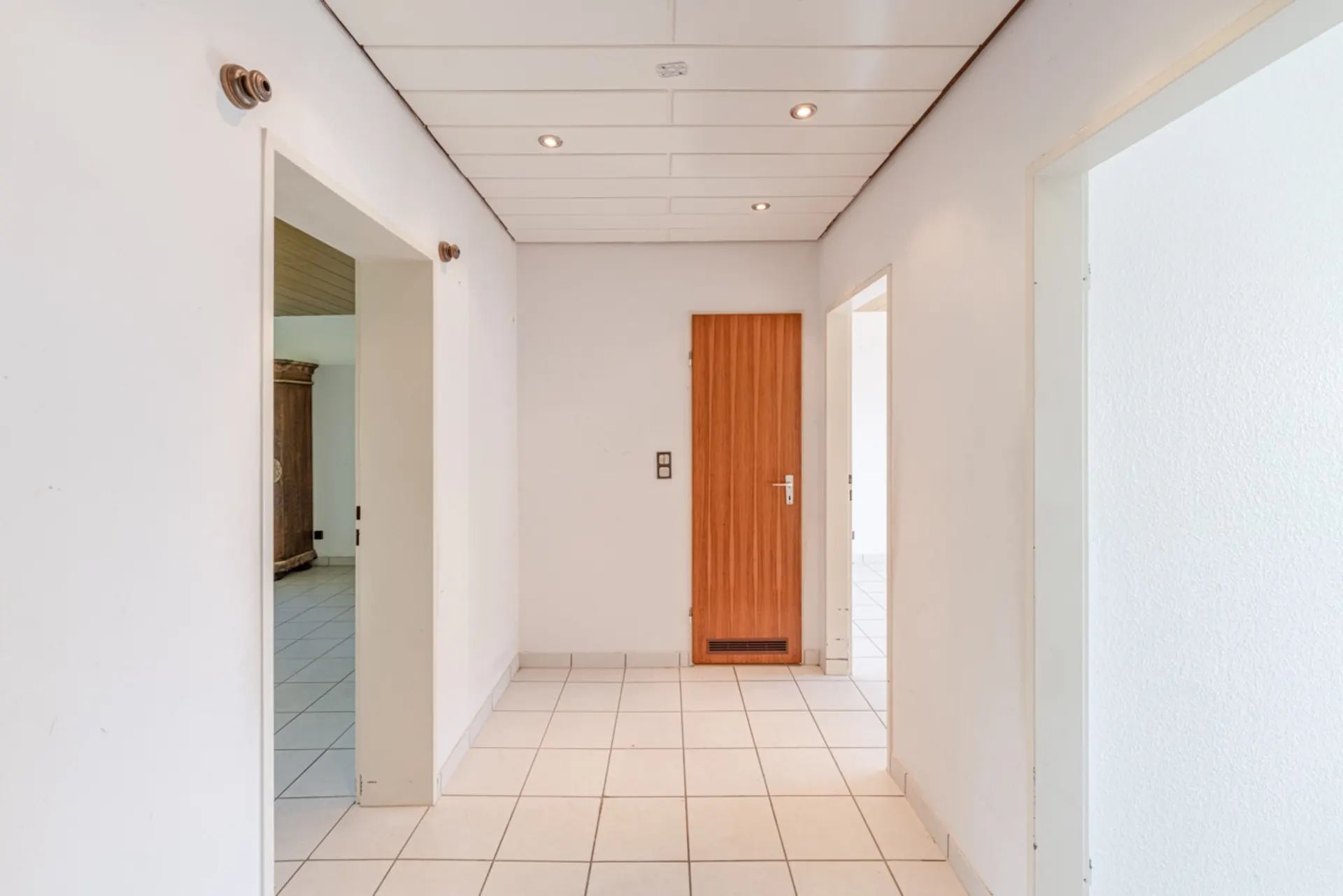 Heller Flur mit weißen Wänden, weißen Fliesen und Holztür in einer Wohnung.