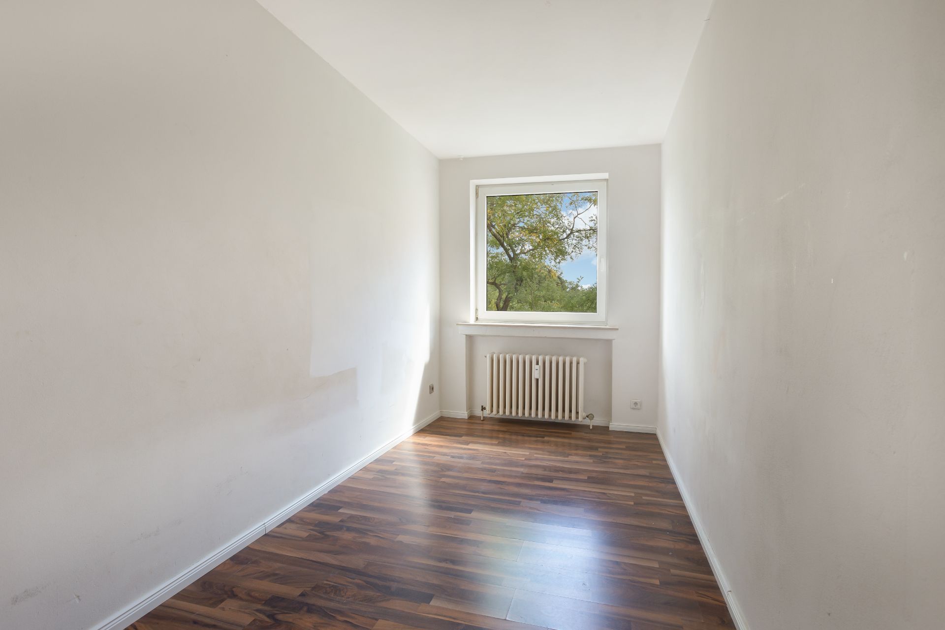 Leerer, schmaler Raum mit Holzfußboden, Fenster und Heizkörper unter dem Fenster.